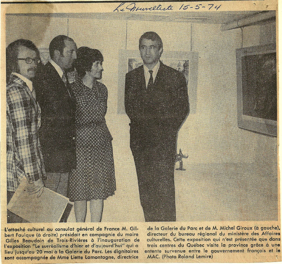 Une photographie en noir et blanc montrant cinq personnes lors de l'inauguration d'une exposition. Un homme en costume est présent, ainsi que quatre autres personnes écoutant ou regardant. La scène semble se dérouler dans une galerie ou un espace d'exposition, avec un mur et une télévision en fond.