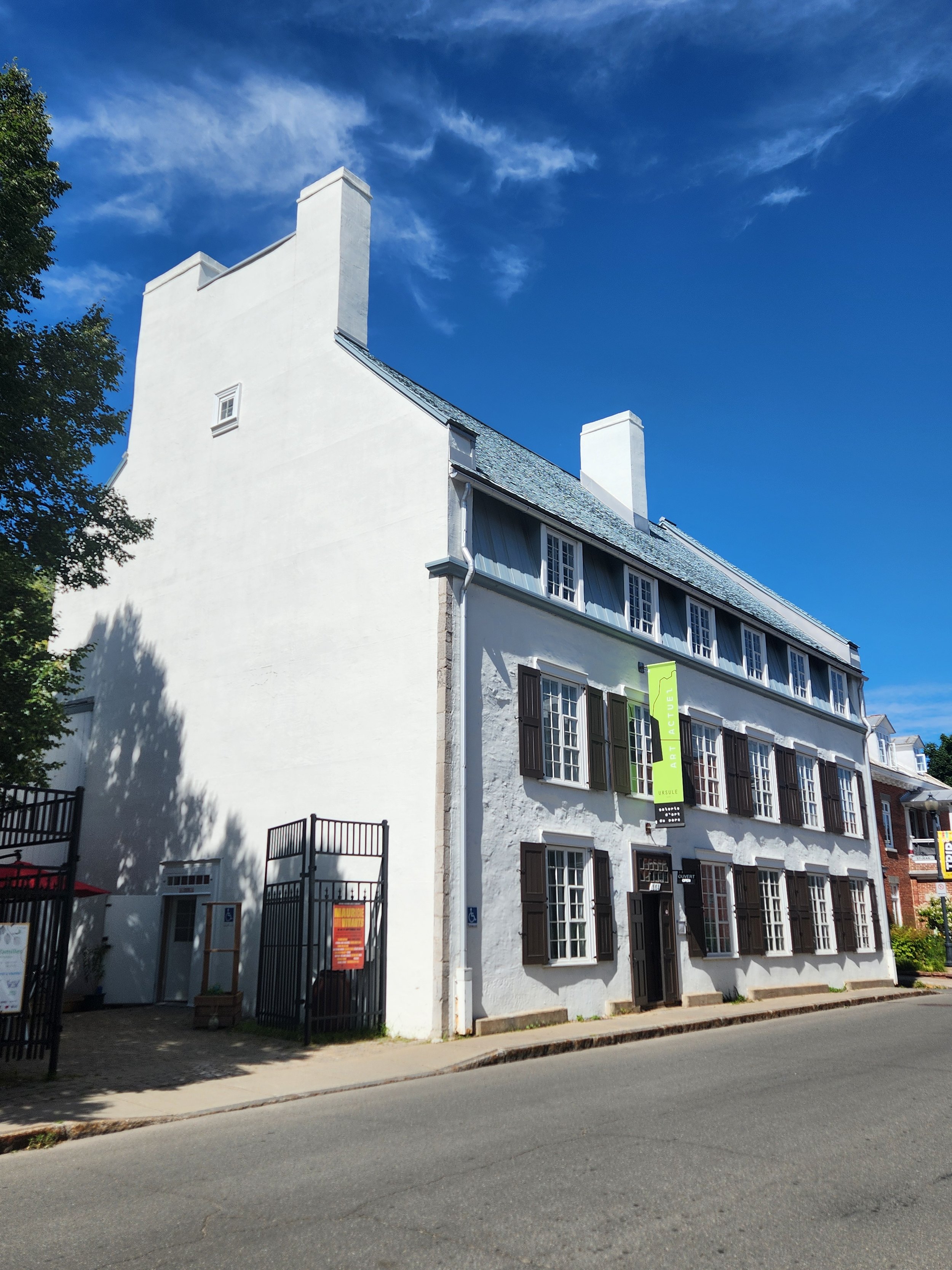 Immeuble blanc avec plusieurs fenêtres à volets en bois, enseigne d'une galerie d'art et un ciel bleu avec quelques nuages.