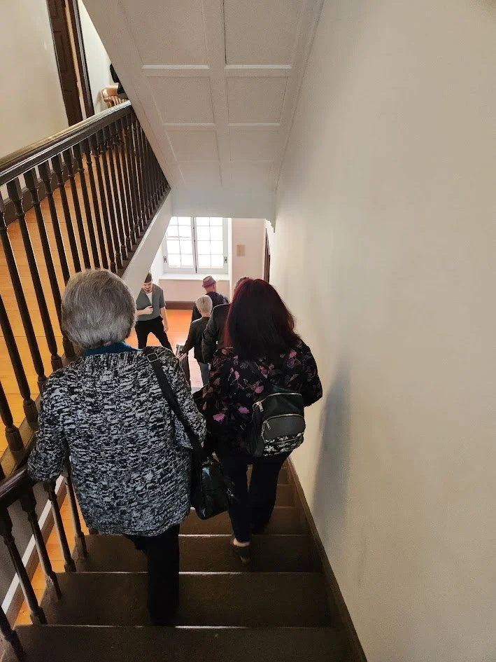 Des personnes descendent un escalier en intérieur dans un bâtiment, avec une grande fenêtre à l'arrière illuminant la scène.