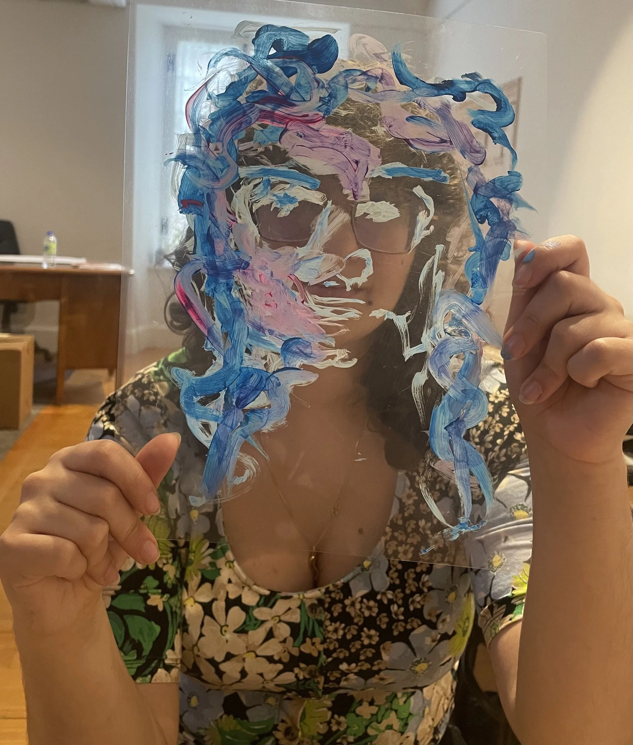 Une femme tient une plaque de verre avec une peinture abstraite en utilisant des couleurs comme le bleu, le rose, le violet et le blanc. Elle porte une robe à motif floral et porte des lunettes. L'arrière-plan montre un intérieur domestique.