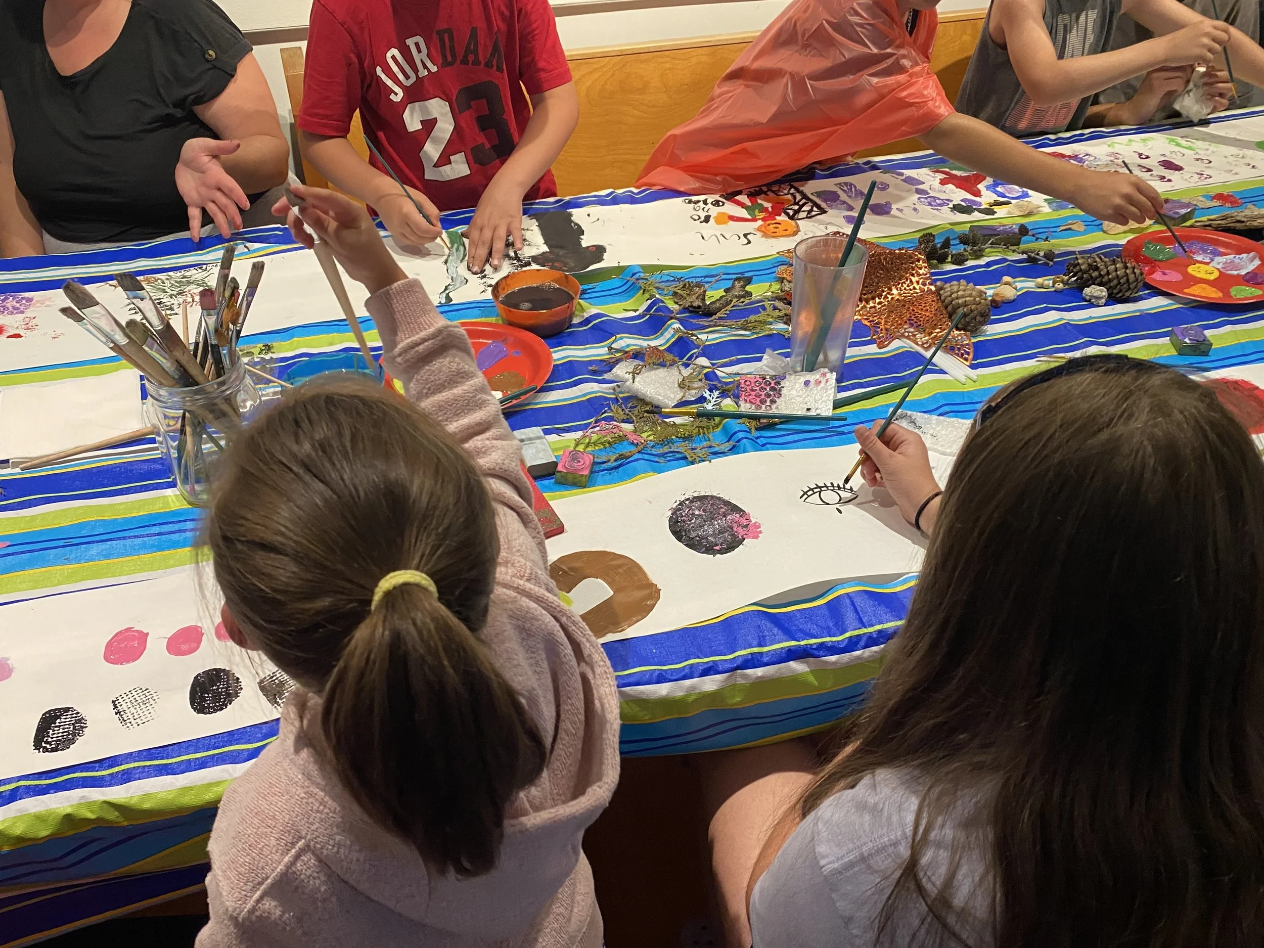 Des enfants et des adultes peignant et décorant des feuilles blanches avec des peintures et divers objets sur une table recouverte d'une nappe colorée.