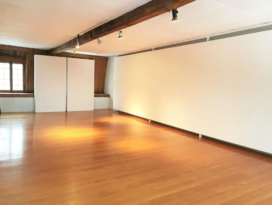 Grand espace vide avec parquet en bois, murs blanc, grande fenêtre en bois et plafond avec poutres en bois et spots d'éclairage.