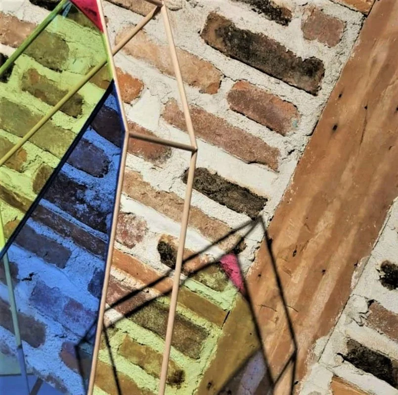 Une structure métallique colorée en zigzag colorée en vert, jaune, bleu et rose posée contre un mur en briques.