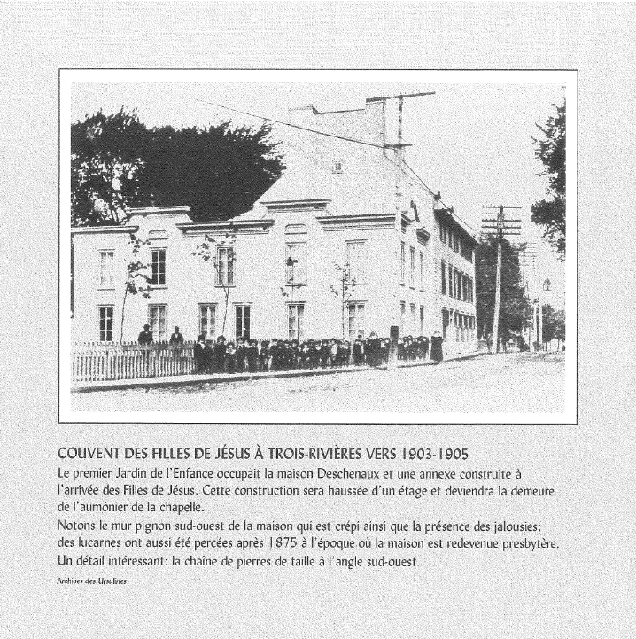 Une vieille photographie en noir et blanc représentant une maison ancienne avec une façade en pierre, entourée d'une clôture en bois, et un groupe de personnes debout devant. Il y a aussi des arbres à l'arrière-plan et des fils électriques au-dessus.