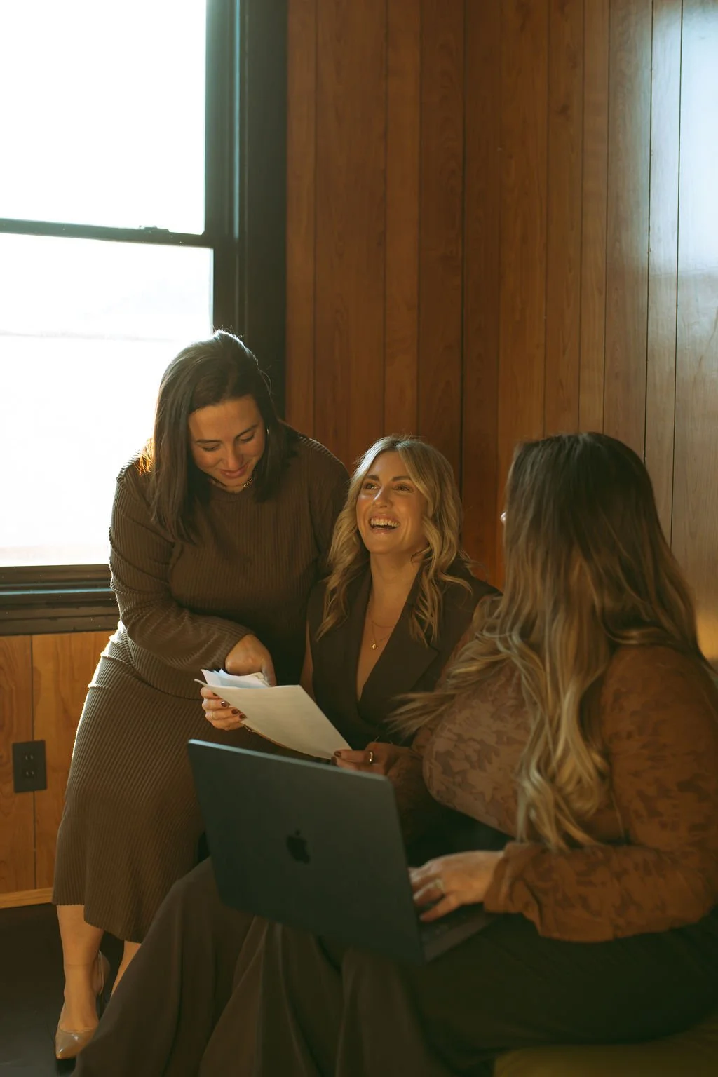 Three women laughing and talking in a room with wooden walls; one is holding papers, another has a laptop.