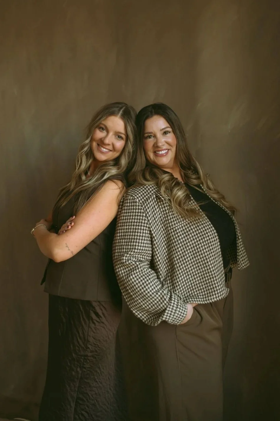 Two women standing back-to-back, smiling, with a brown background. One woman has long, light brown hair, and the other has long, dark brown hair. They are dressed in smart casual attire.
