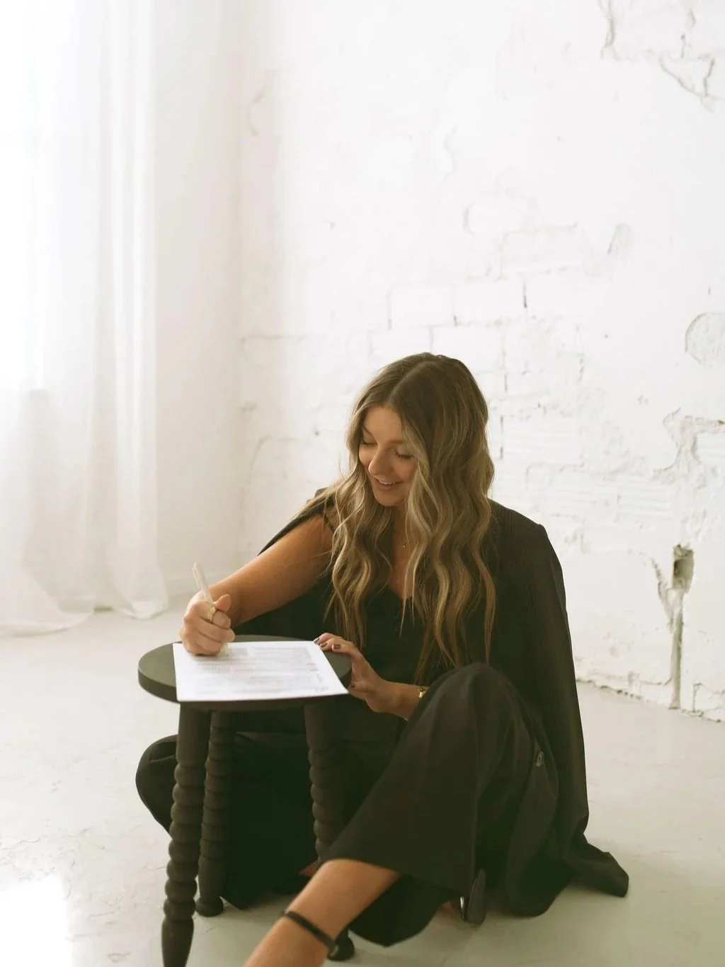 A woman with long wavy hair sitting on the floor, smiling, as she writes on a paper placed on a small black side table in a bright room with white brick walls and a window with white curtains.