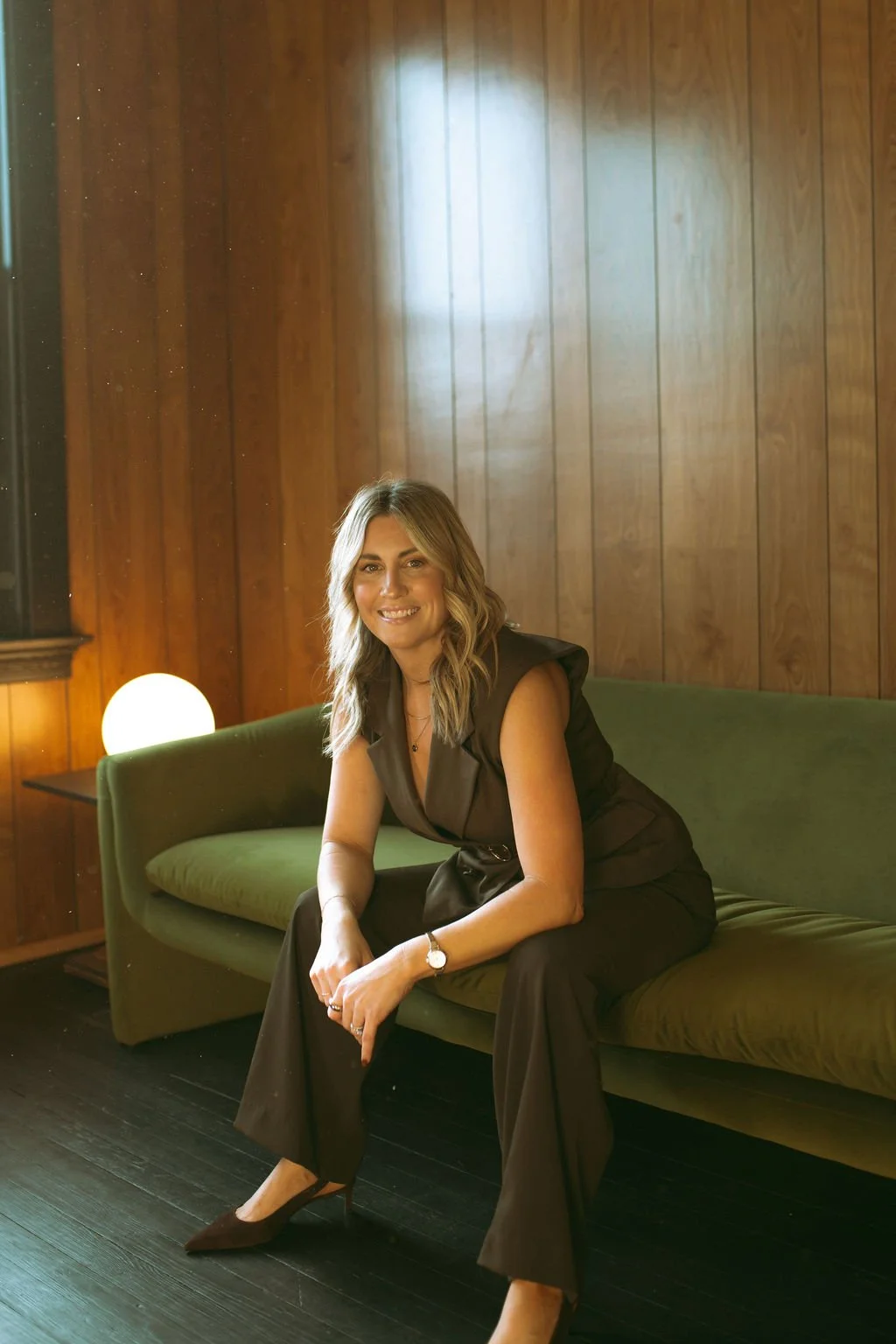 A woman sitting on a green sofa in a room with wood-paneled walls, smiling at the camera, wearing a black sleeveless jumpsuit and brown high heels.