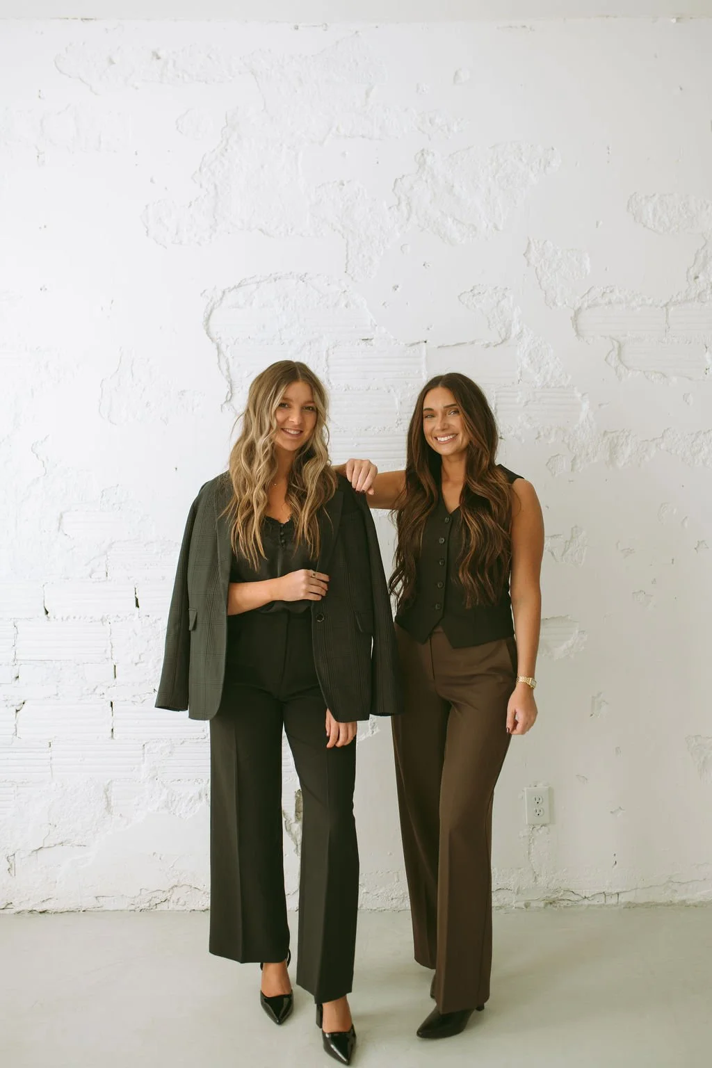 Two women standing side by side smiling at the camera against a white brick wall. One woman is holding a gray blazer, and the other has her arm around her friend's shoulder.