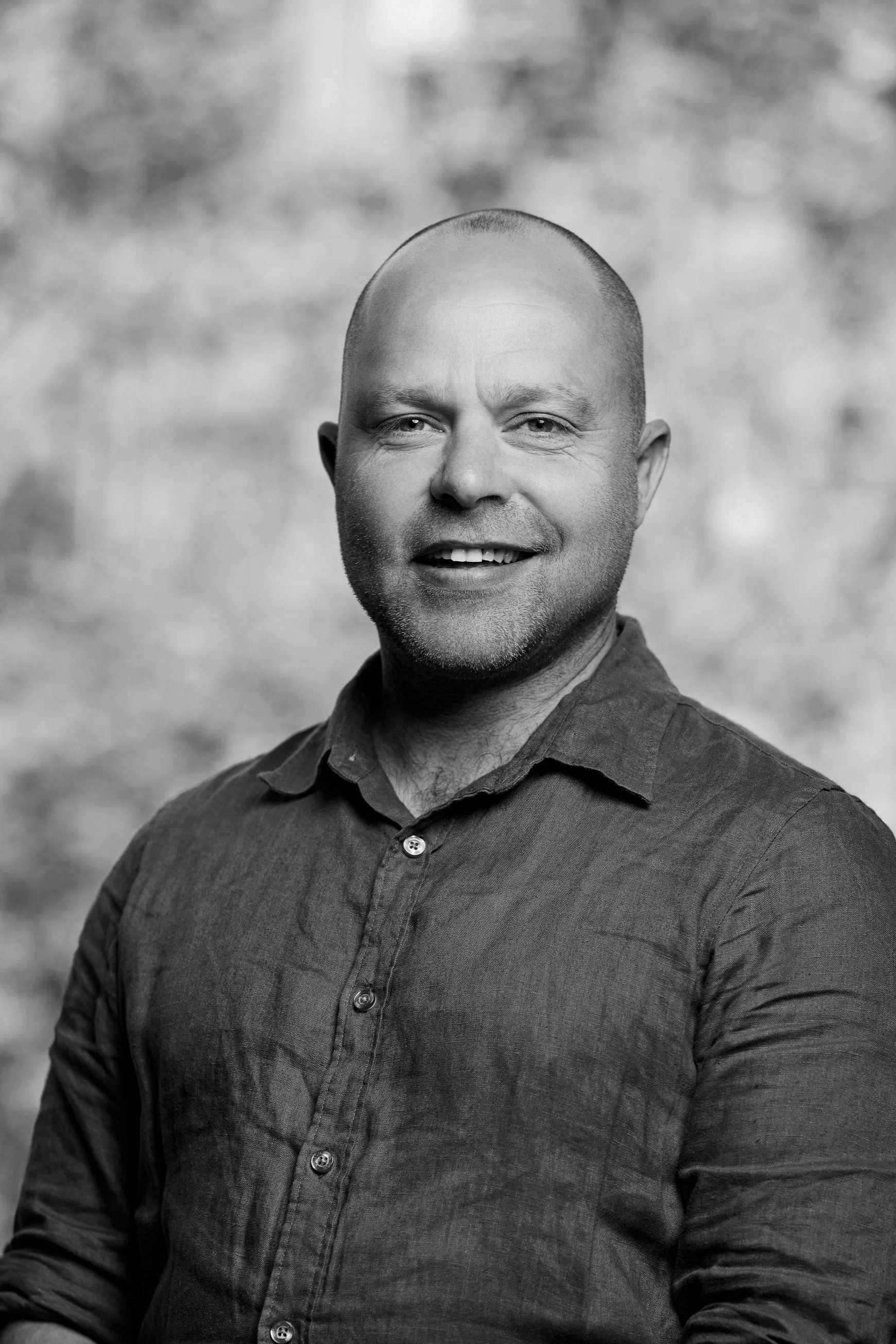 Black and white professional portrait of a smiling bald man wearing a button-up shirt against a blurred background.