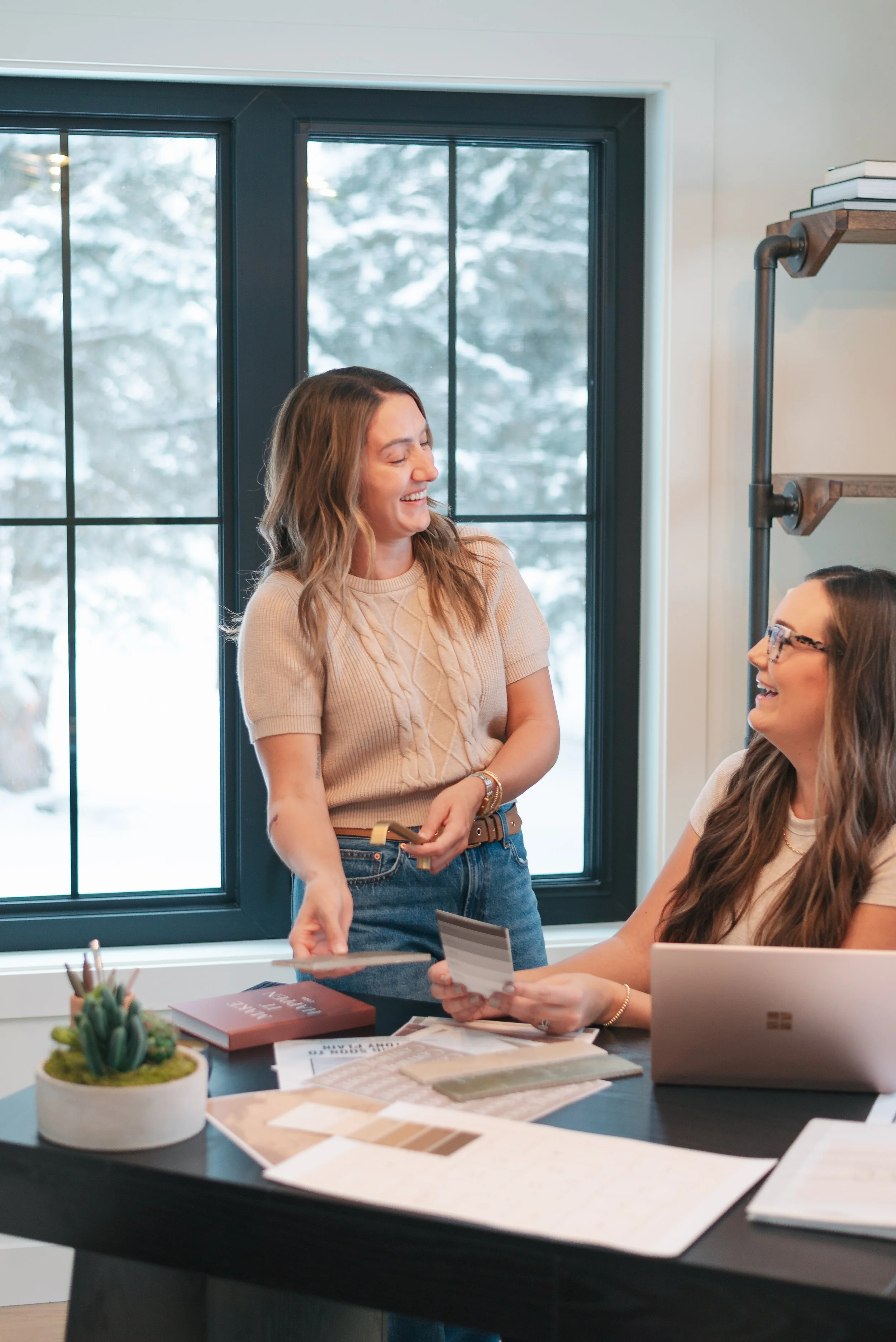 Two women in an office, one standing and smiling, the other sitting and holding a color swatch, with a laptop and design samples on the table, large window with a snowy landscape outside
