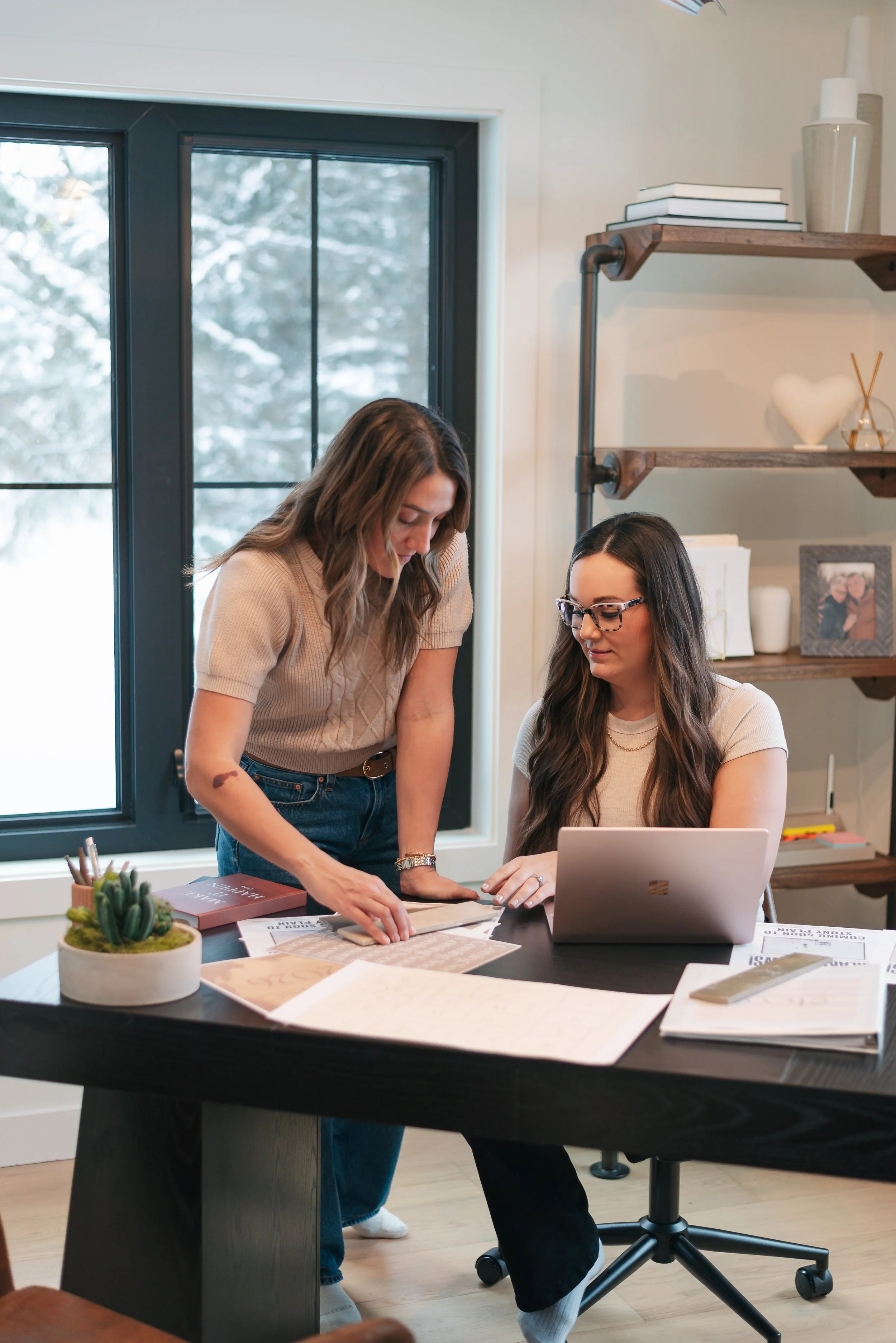 Two women working together at a desk in a home office with a window showing snow outside. One woman is sitting with a laptop, and the other is standing and pointing at documents.