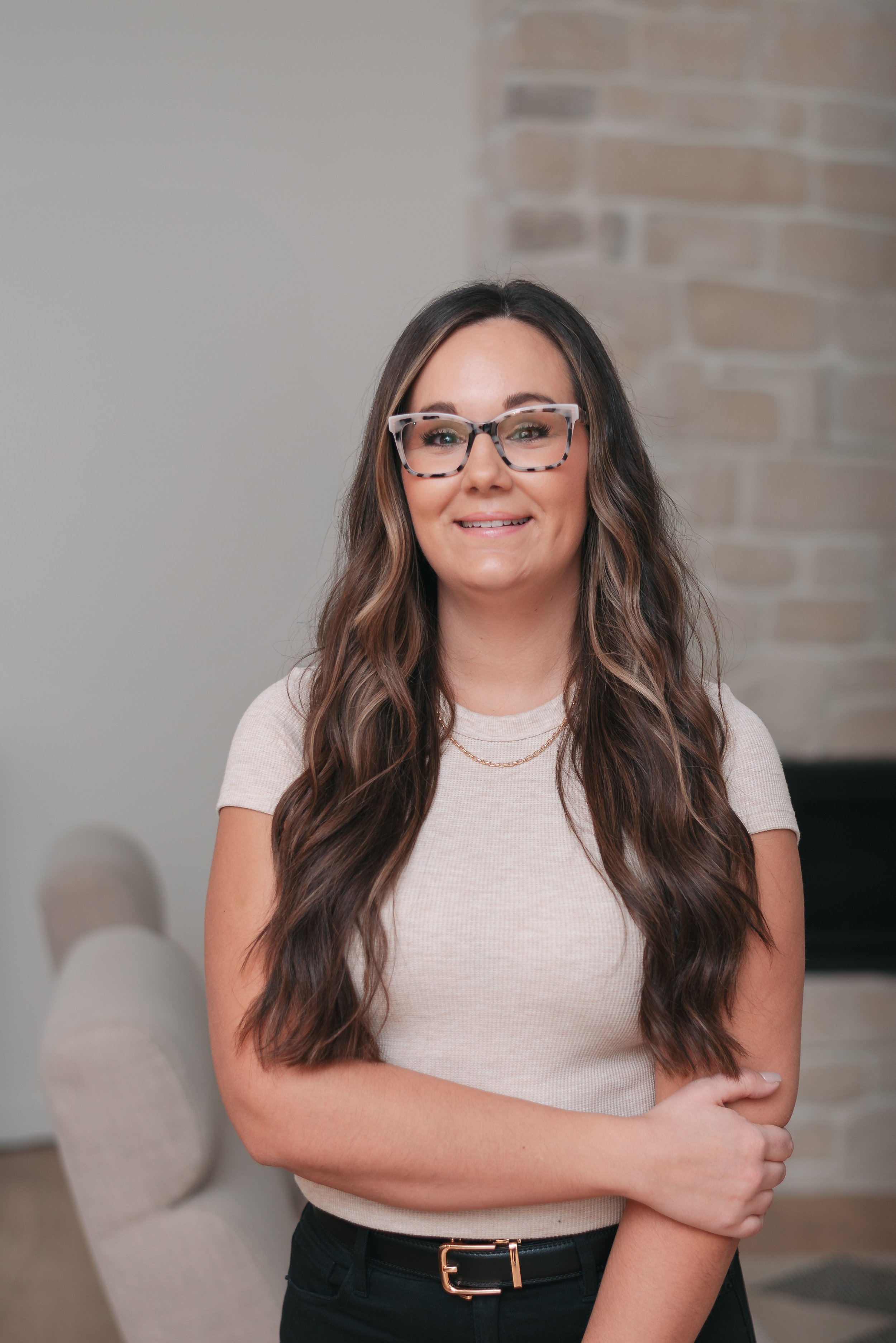 A woman with long, wavy brown hair wearing glasses, a beige short-sleeve shirt, and black pants, standing indoors in front of a stone fireplace and a light-colored wall.