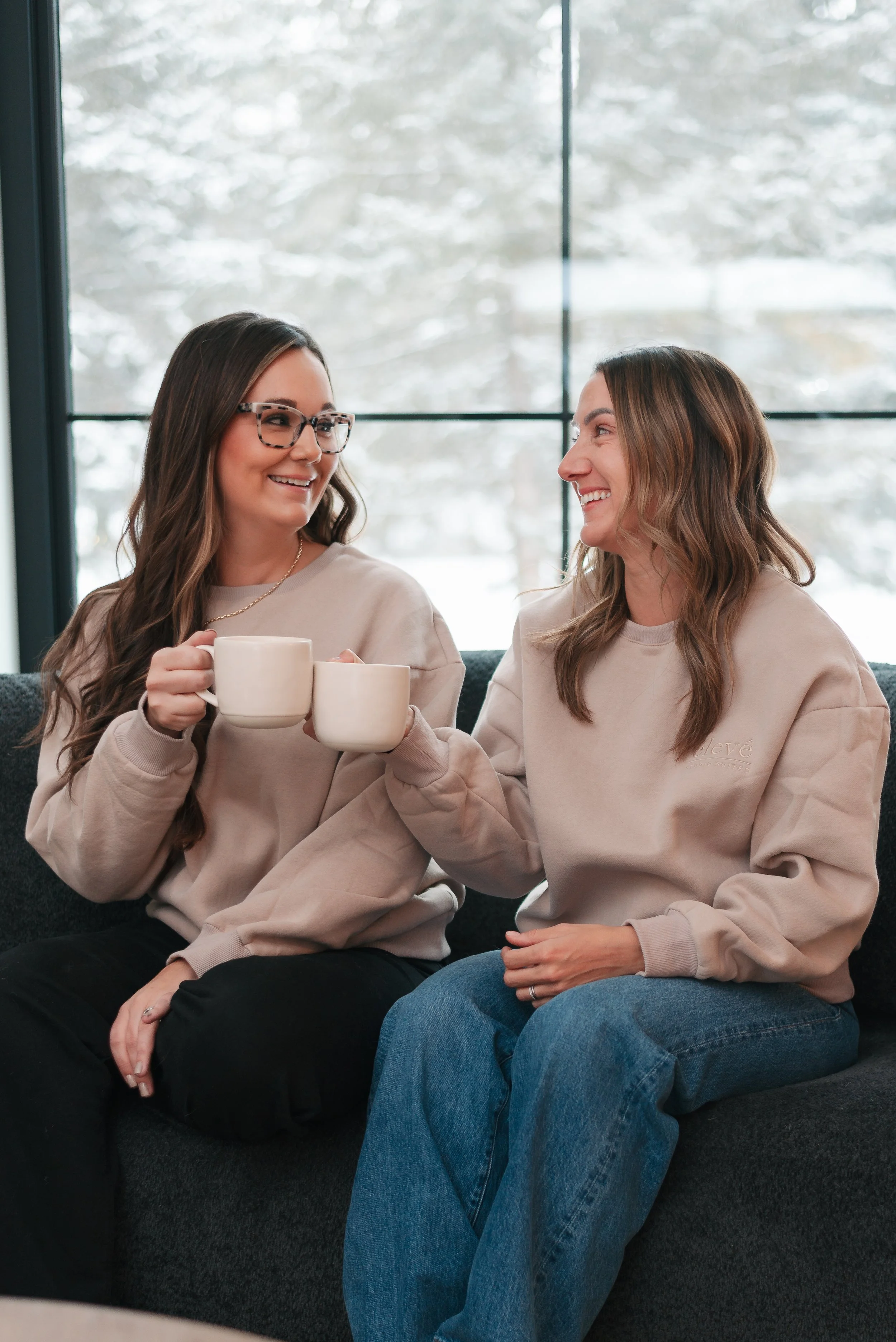 Two women sitting on a dark gray sofa, smiling and toasting with white mugs, with snow-covered trees visible through large windows behind them in Stony Plain, Alberta.