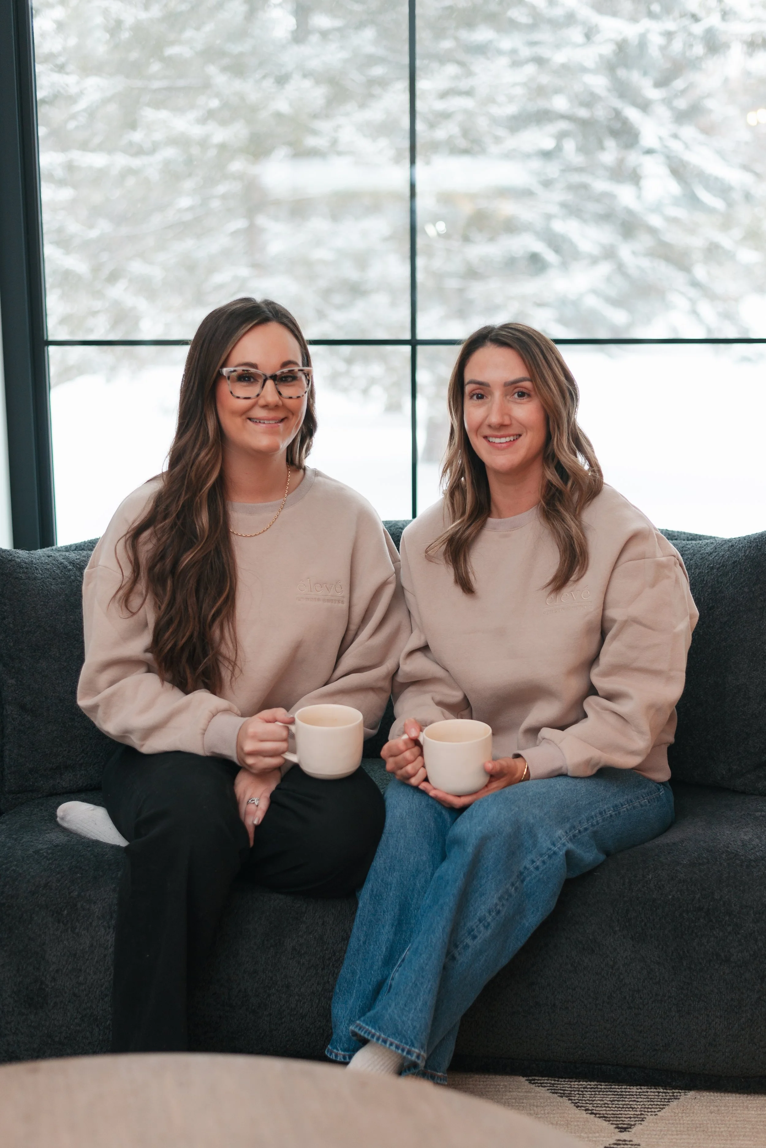 Two women sitting on a dark gray couch inside, holding white mugs, wearing beige sweatshirts, with snow-covered trees visible through a large window behind them.