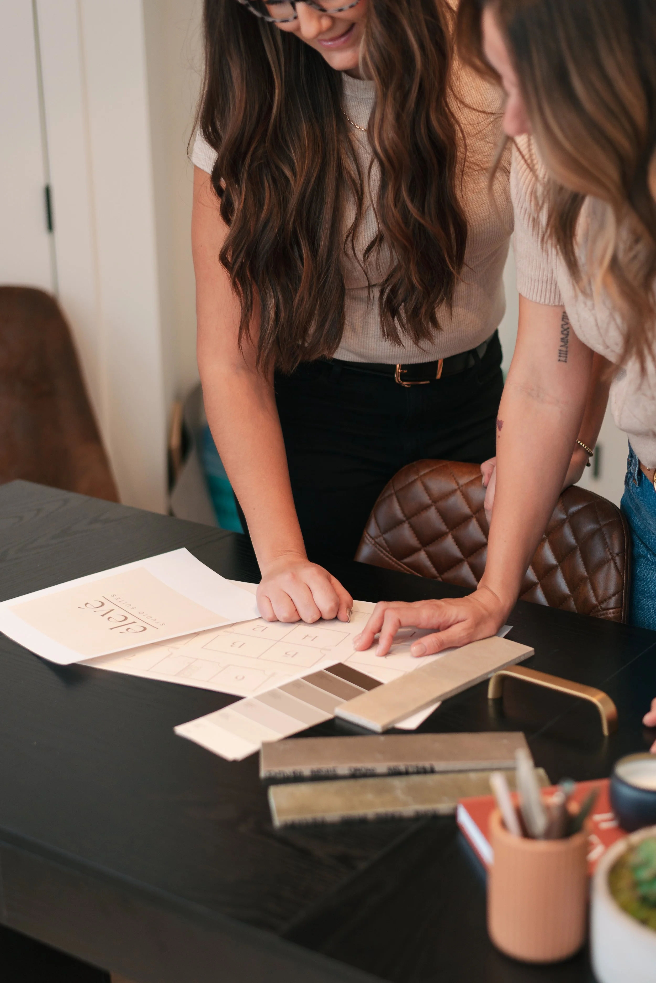 Two women are standing over a table discussing interior design, with samples of paint colors, fabric swatches, and design plans on the table.
