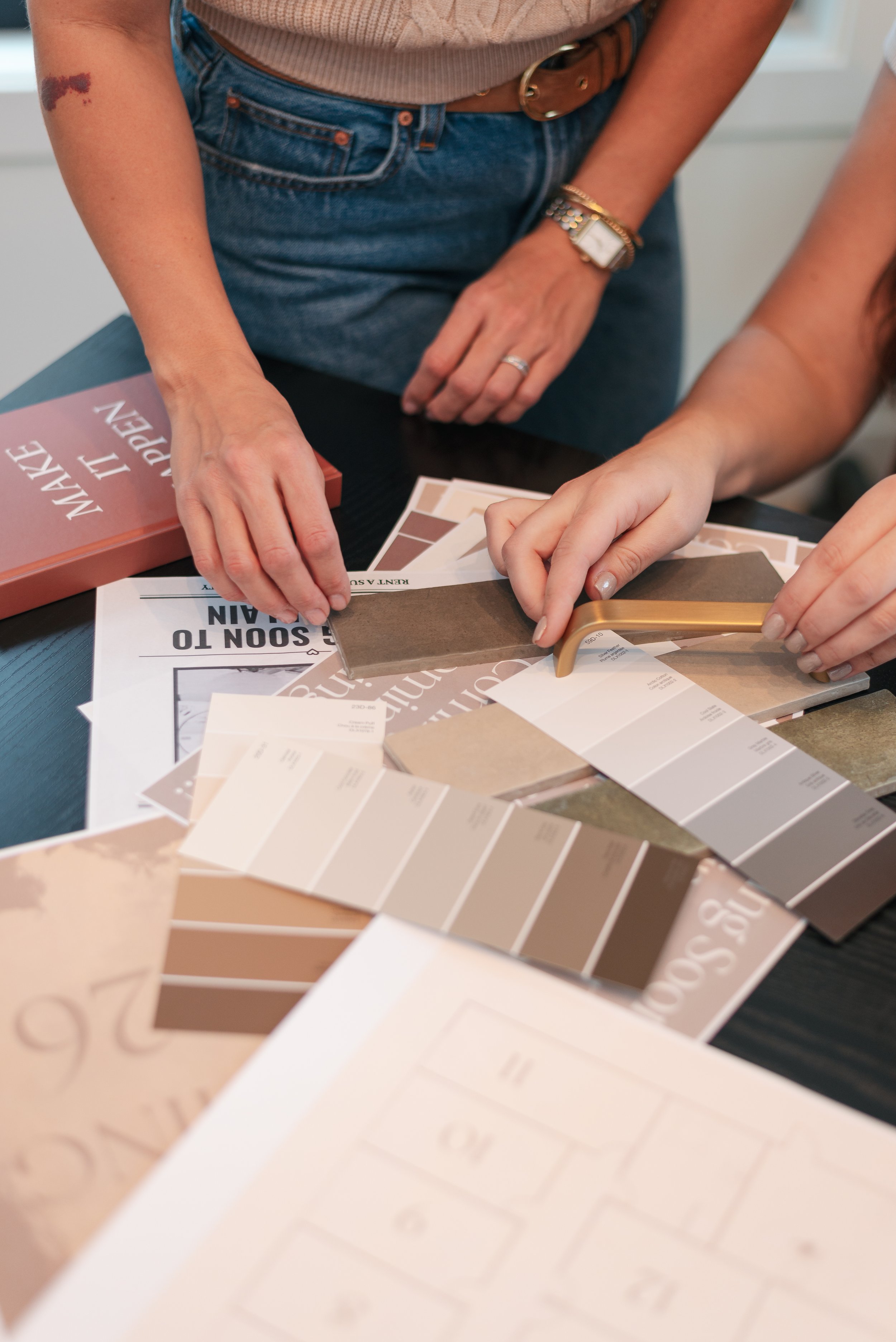 Two women are examining and selecting neutral-colored paint swatches and samples on a black table, with a 'Make It Repe' book also on the table.