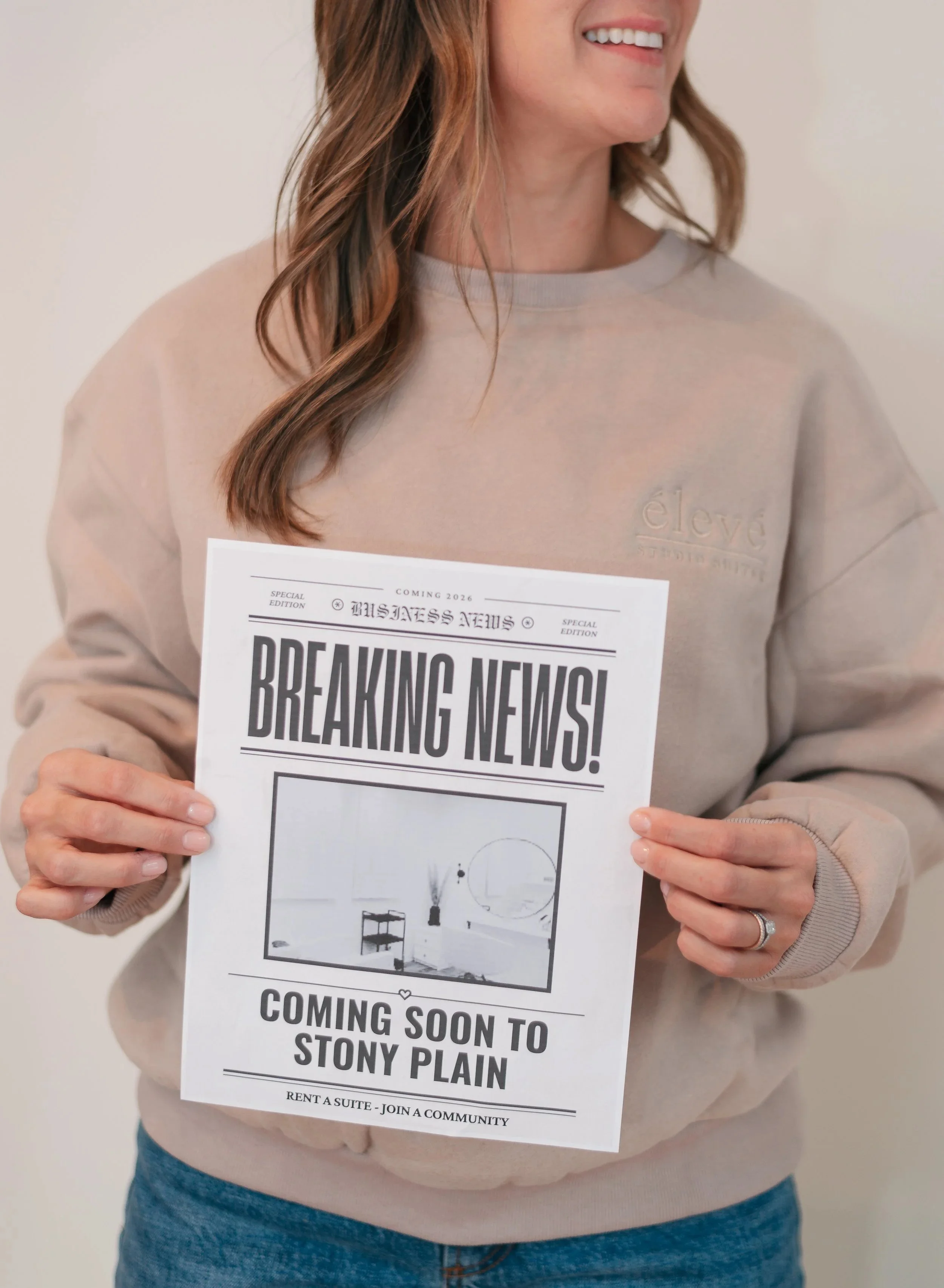 A woman holding a flyer that says 'Breaking News! Coming Soon to Stony Plain.' The flyer features a minimalist interior image and indicates potential real estate or community news.