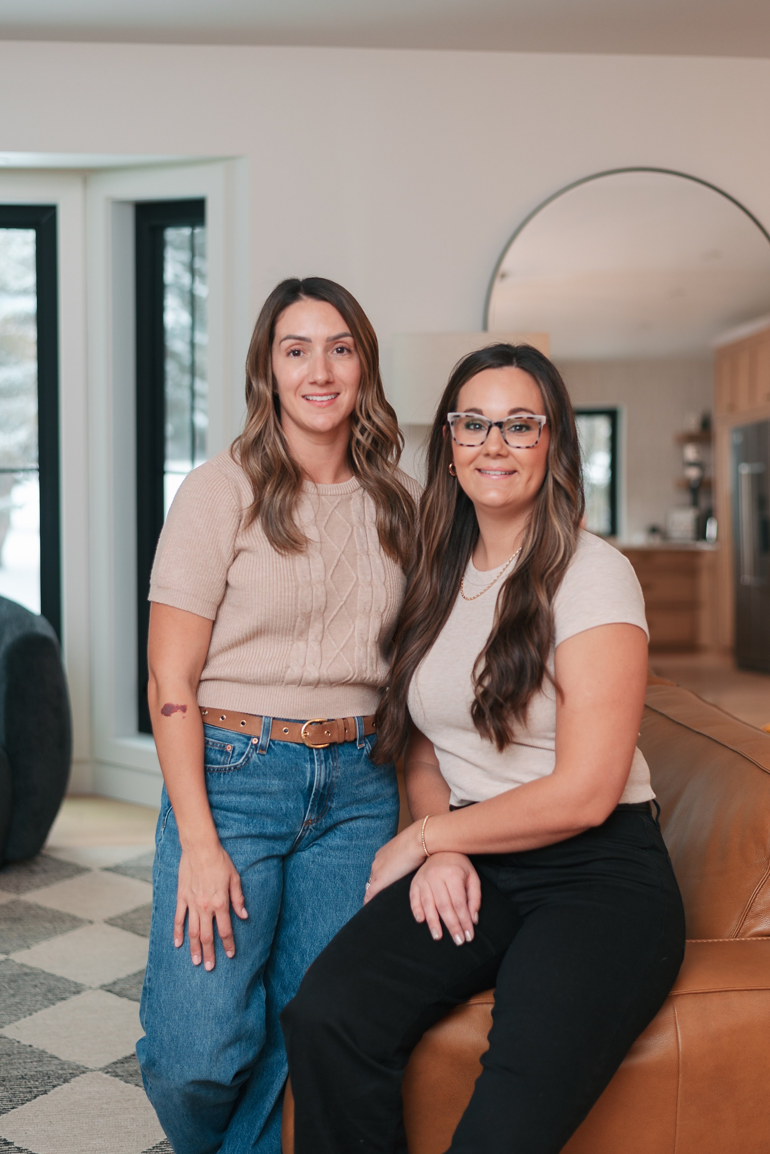 Two women with long wavy hair smiling in a cozy living room, one standing and the other sitting on a leather couch, with large windows showing a winter scene outside.