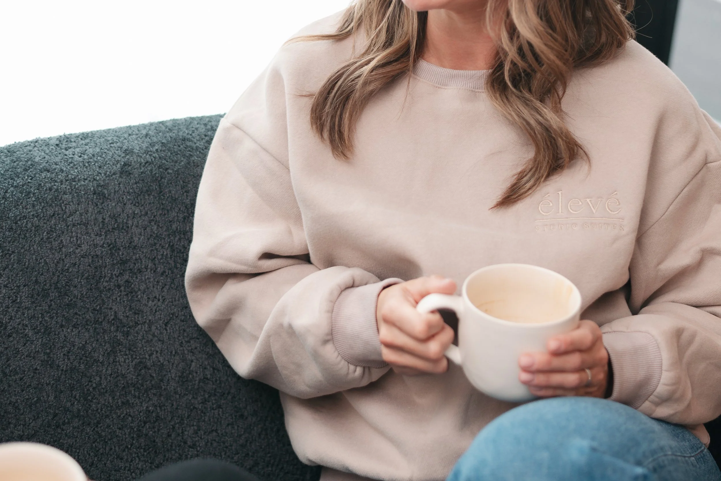 A woman in a beige sweatshirt sitting on a gray couch, holding a white mug, with orange hair, and blue jeans.