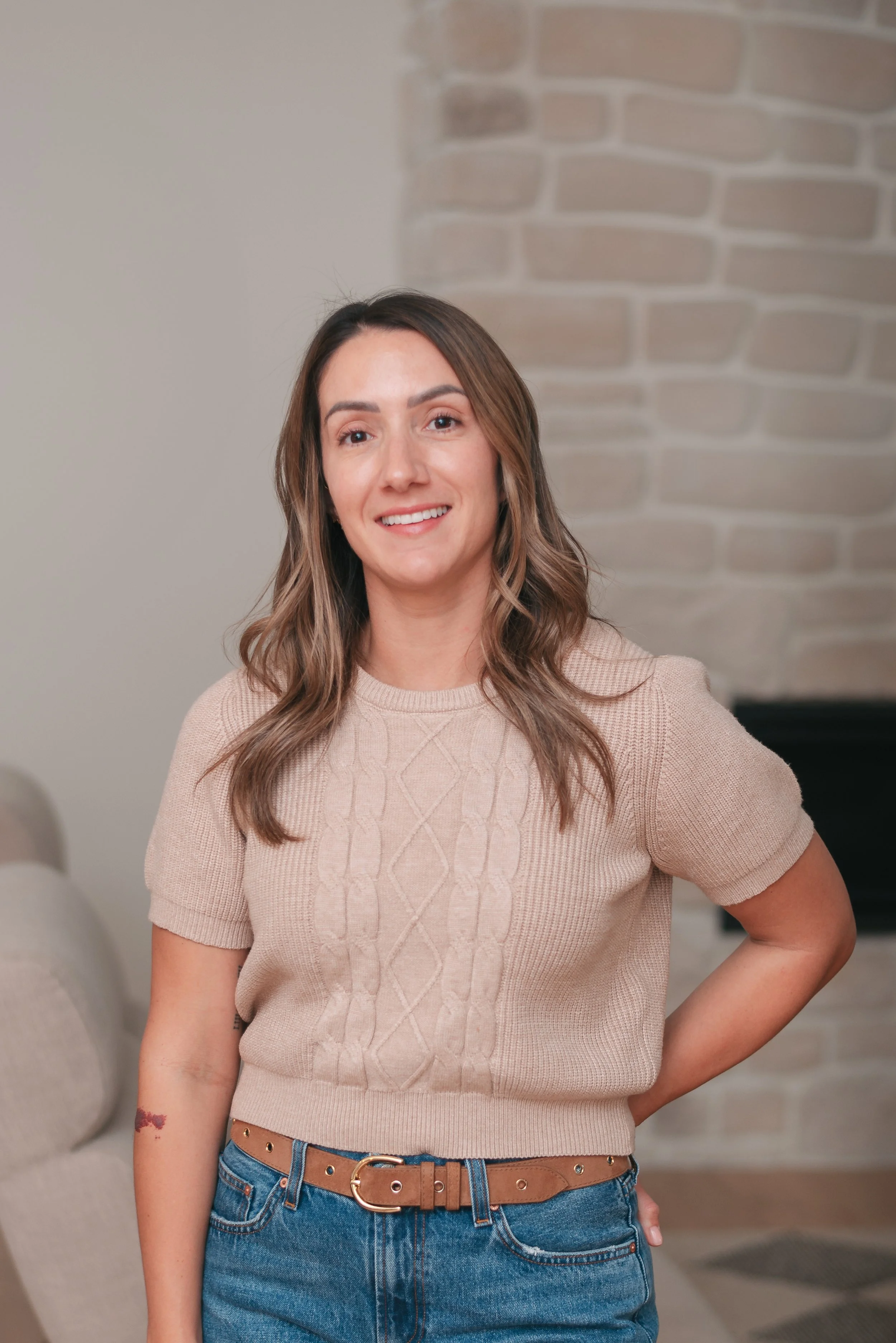 A woman wearing a beige knit sweater and blue jeans standing in a room with a brick wall and a beige couch.