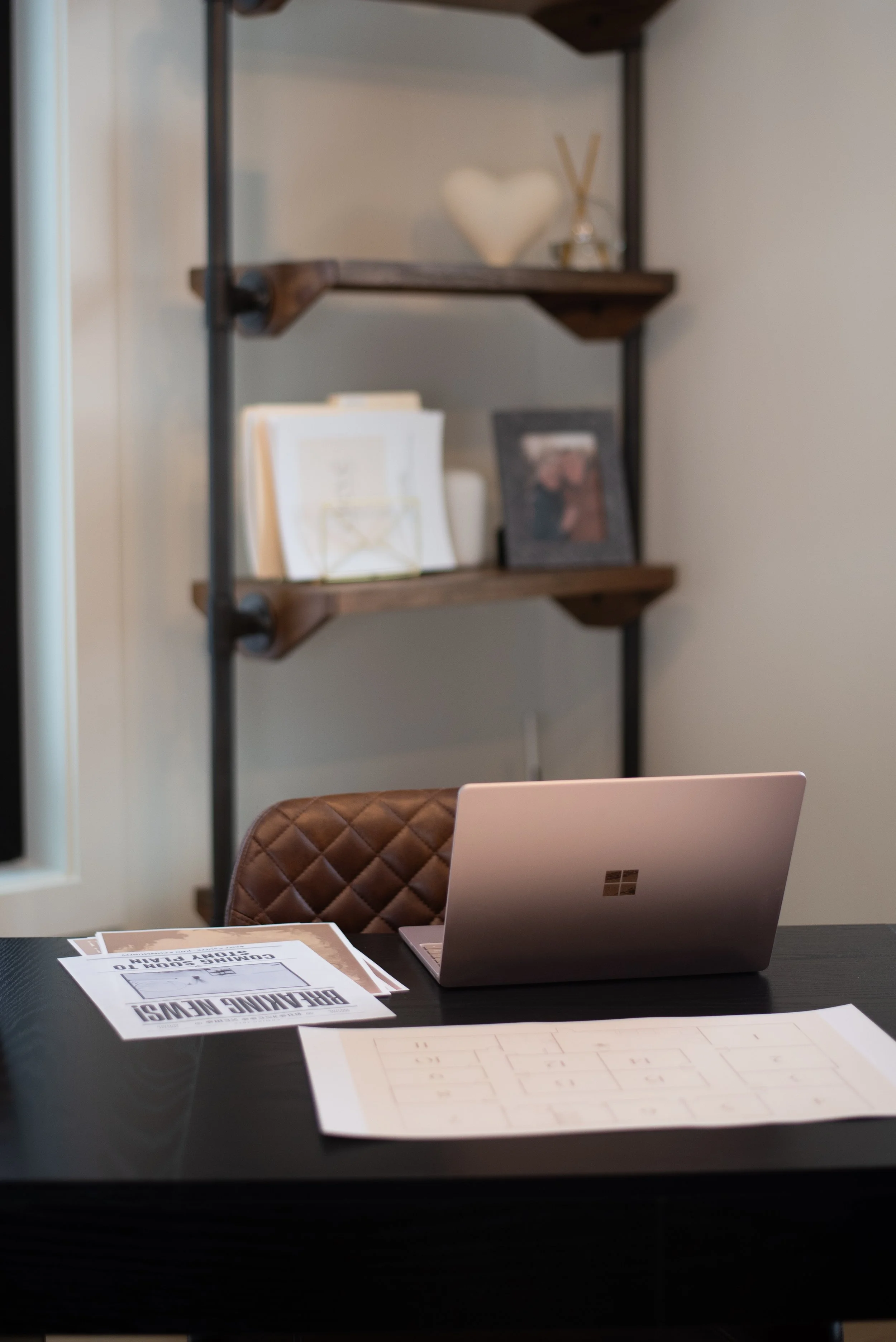 An office or workspace with a black desk, a silver Microsoft Surface laptop, some papers, and a brown quilted chair. In the background, there's a wooden and metal shelf with decorative items and framed photos.