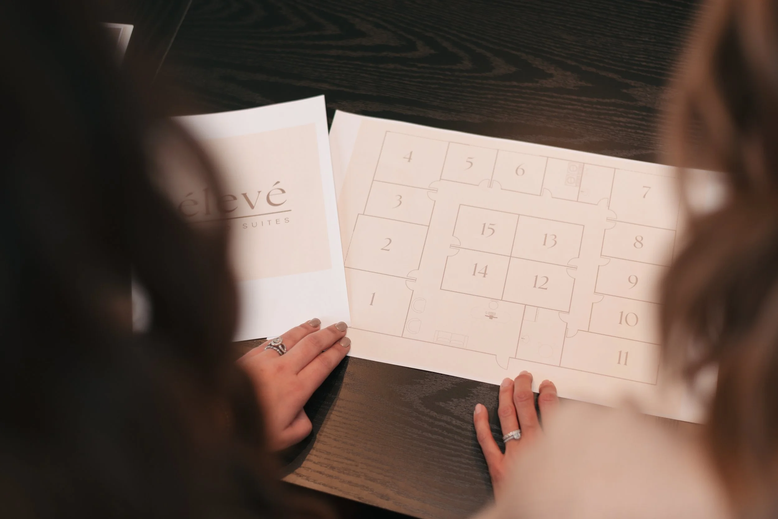 Two women look at a printed hotel floor plan or room layout, with a booklet that says 'élevé' on the cover, on a dark wood table.