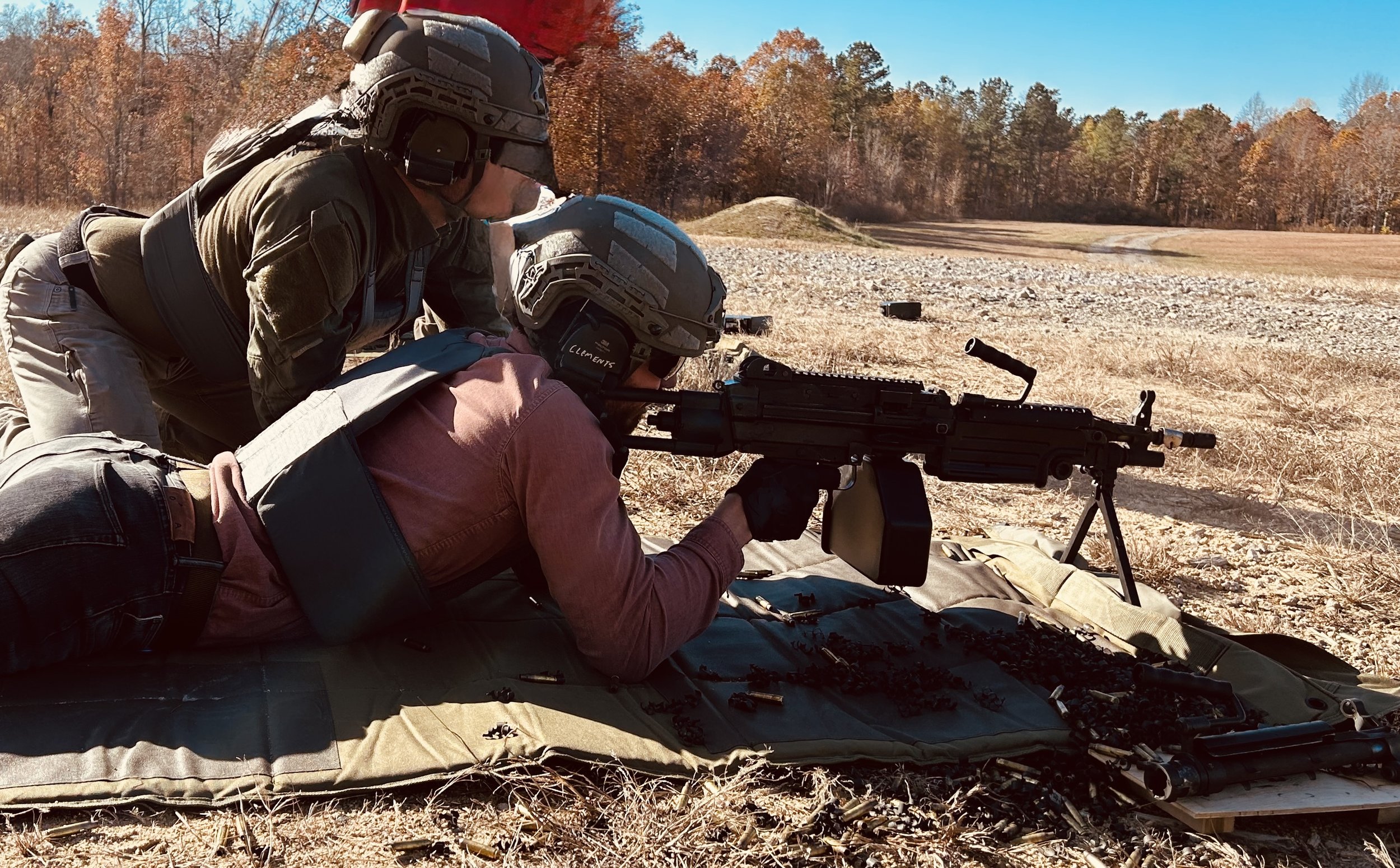 Two individuals in military gear, wearing helmets and protective clothing, are lying on the ground outdoors during the daytime. One is aiming with a mounted machine gun positioned on a bipod, while the other is assisting or observing. The scene is set in an open, rural area with dry grass, dirt, and trees in the background, under clear blue skies.