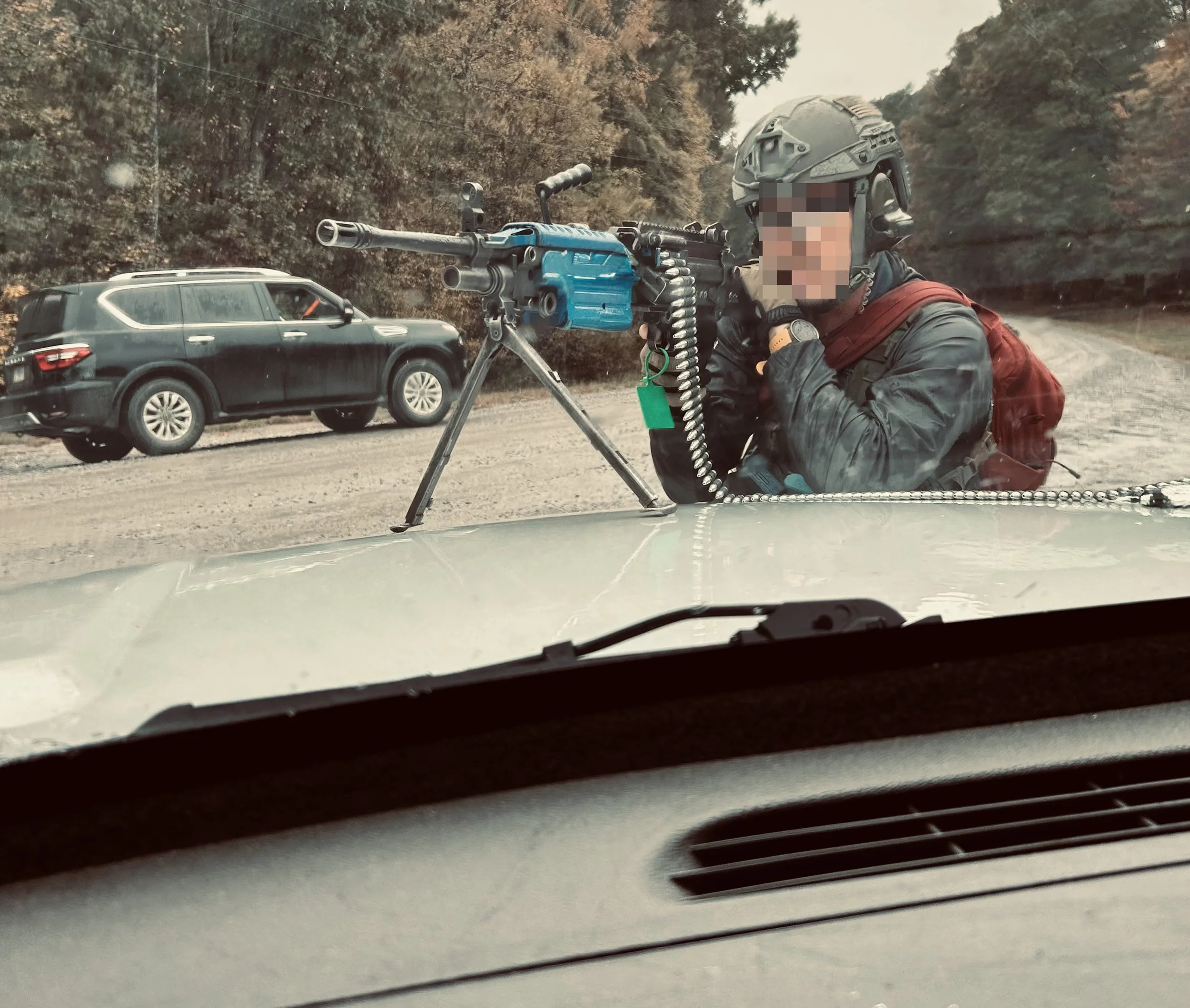 A soldier in tactical gear with a helmet and red backpack operating a mounted machine gun on a vehicle's hood, on a rural road with trees and a black SUV in the background.