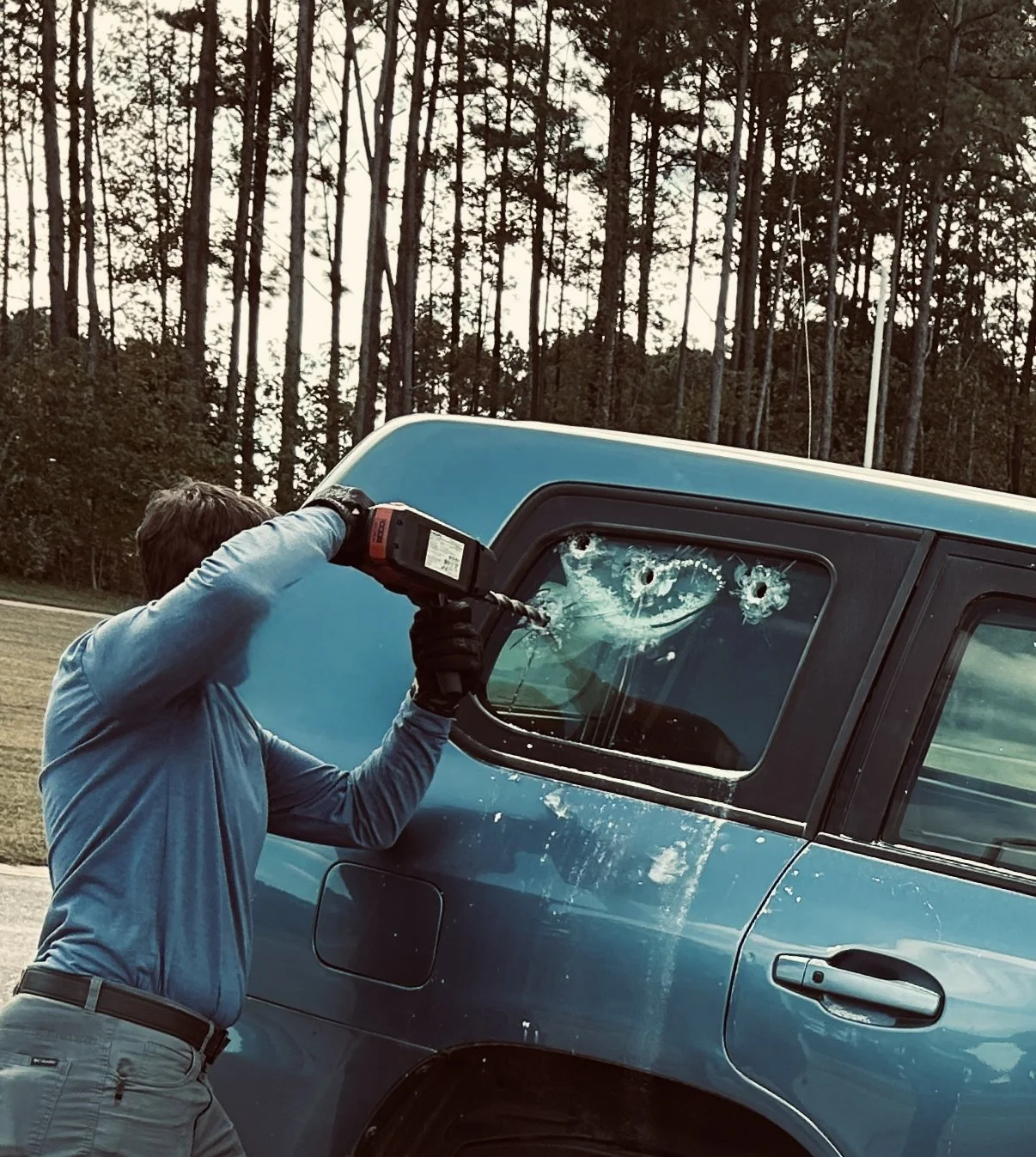 Person using a drill to create bullet holes in a blue truck's window, with a forest in the background.