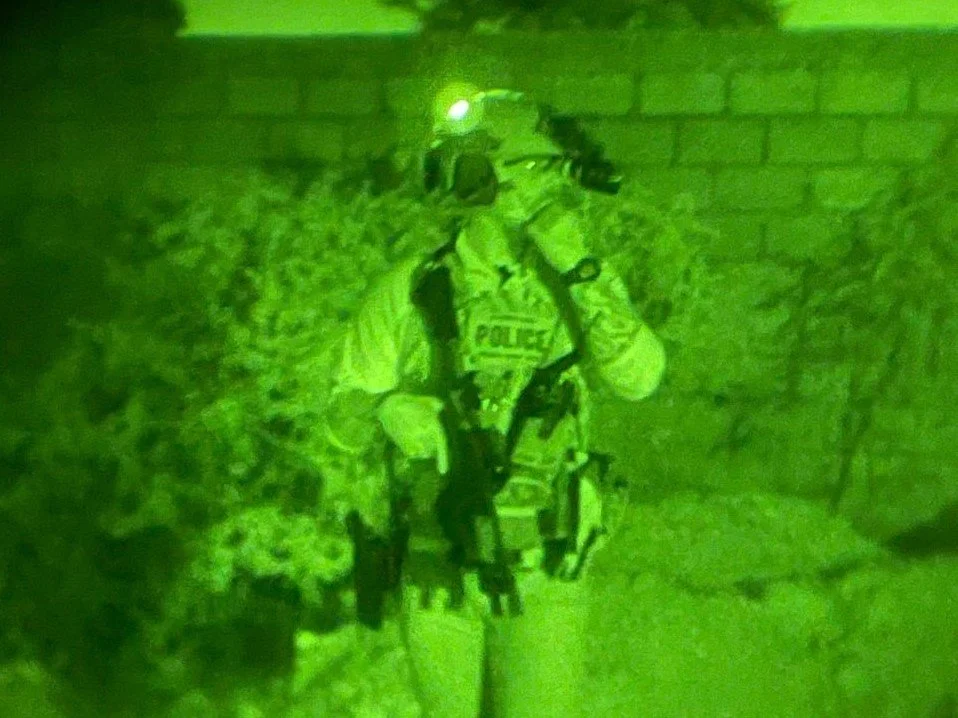 Night vision image of a police officer in tactical gear walking outdoors at night near a brick wall.