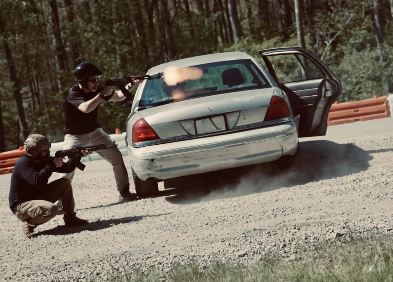 Two men in black shirts and helmets shoot rifles at a car that is crashing into a barrier on a dirt road in a wooded area.