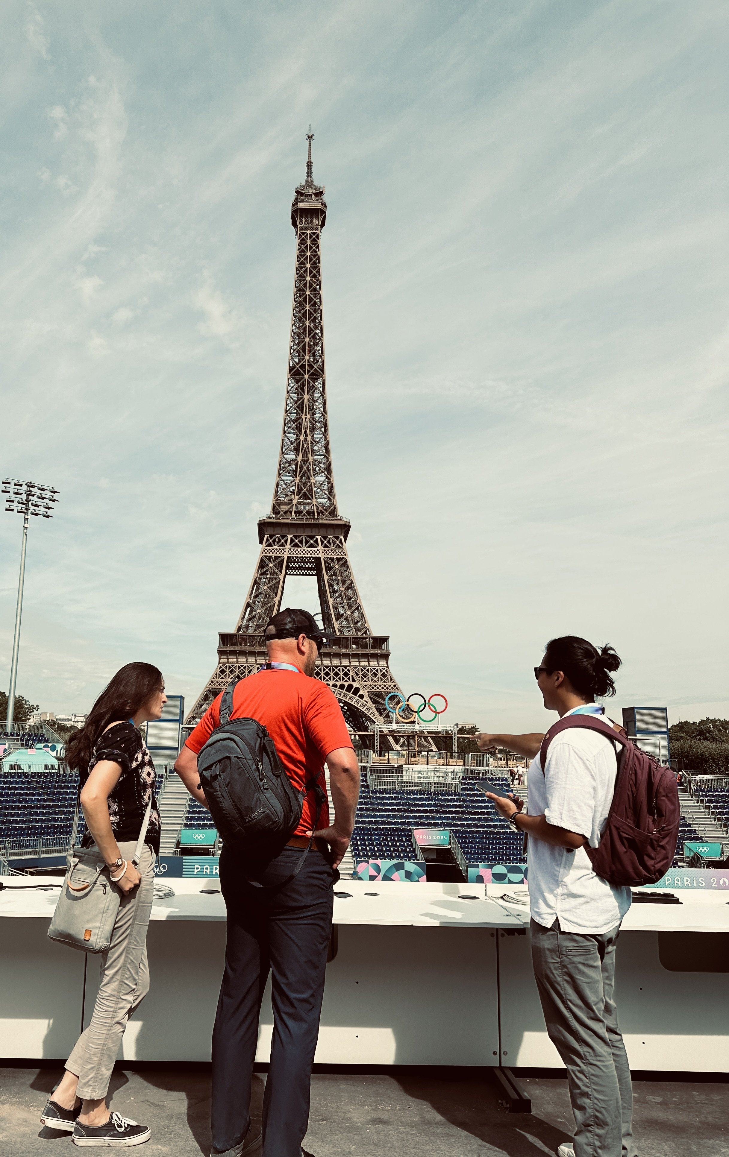 Three people standing in front of the Eiffel Tower in Paris, France, with Olympic rings visible near the tower, engaging in conversation during daytime.