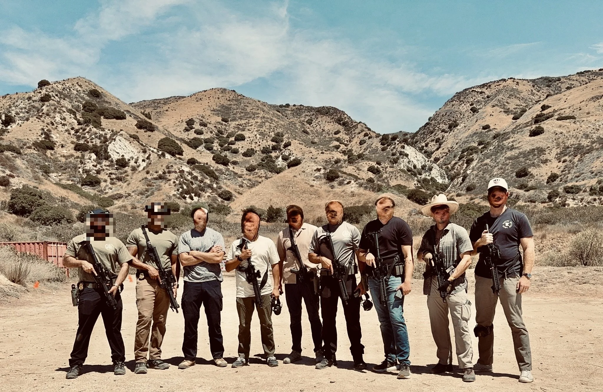 Group of nine individuals standing outdoors in a desert-like area with hills in the background. They are holding rifles and wearing casual clothing, some with hats and tactical gear.