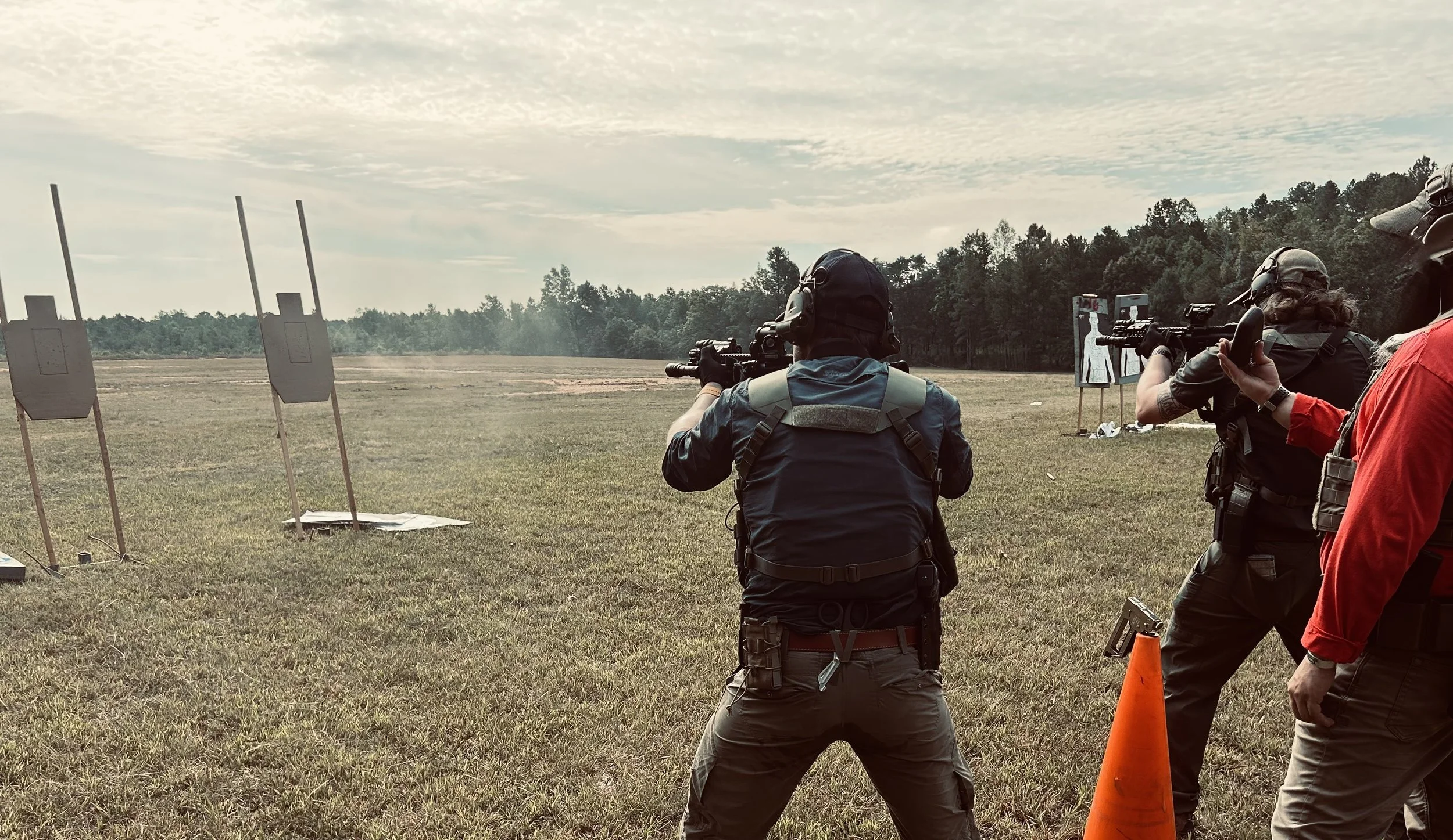 Multiple people participating in a firearms shooting event in an open field, with targets set up in the distance and an instructor or range officer overseeing the activity.