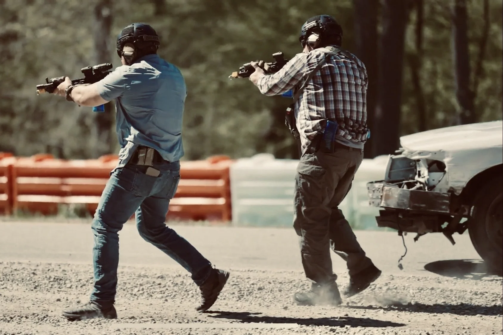 Two men wearing helmets and casual clothing are aiming rifles while walking; one is near a damaged car with a missing front bumper.