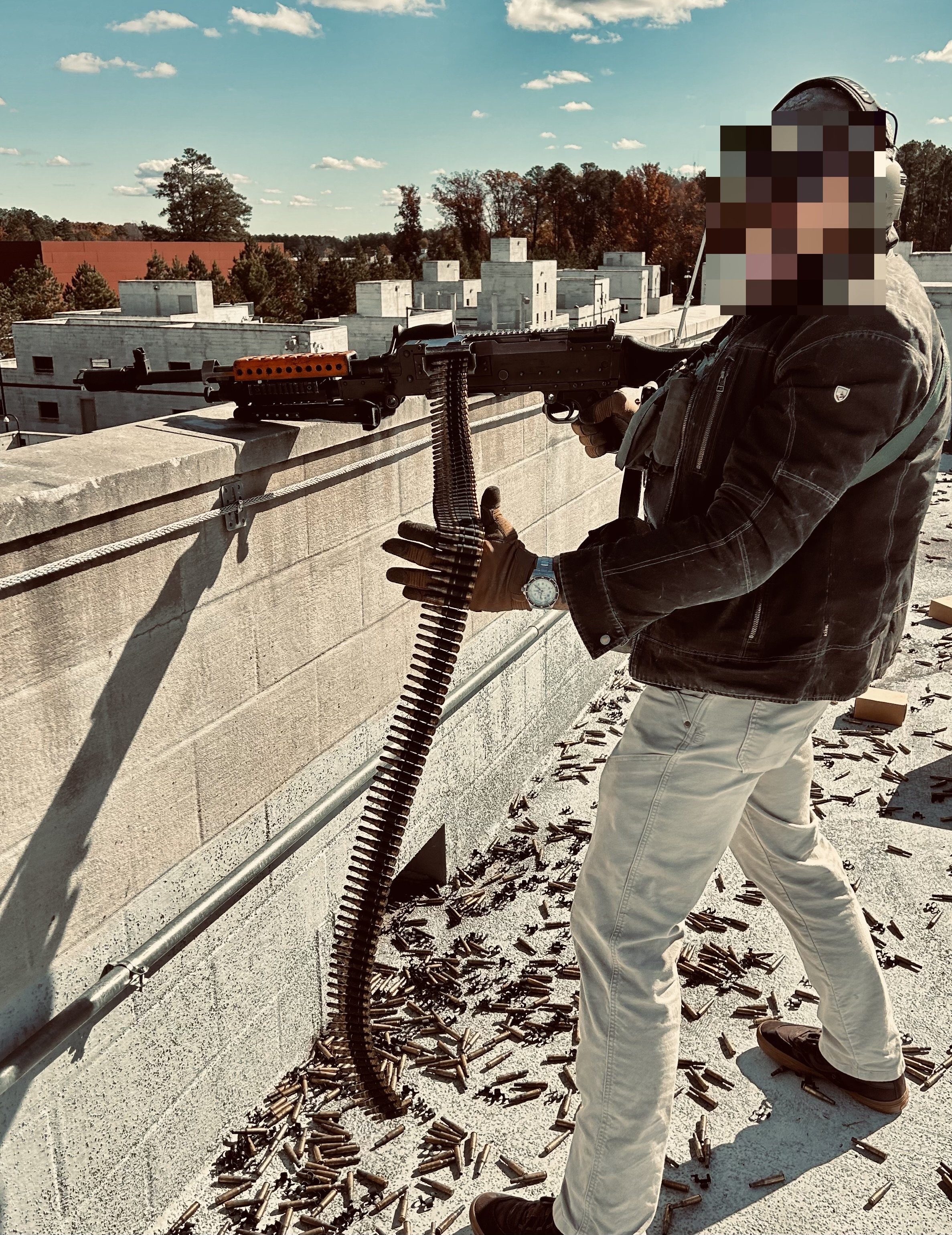 Person firing a rifle on a rooftop, surrounded by spent bullet casings, with a city skyline and trees in the background.