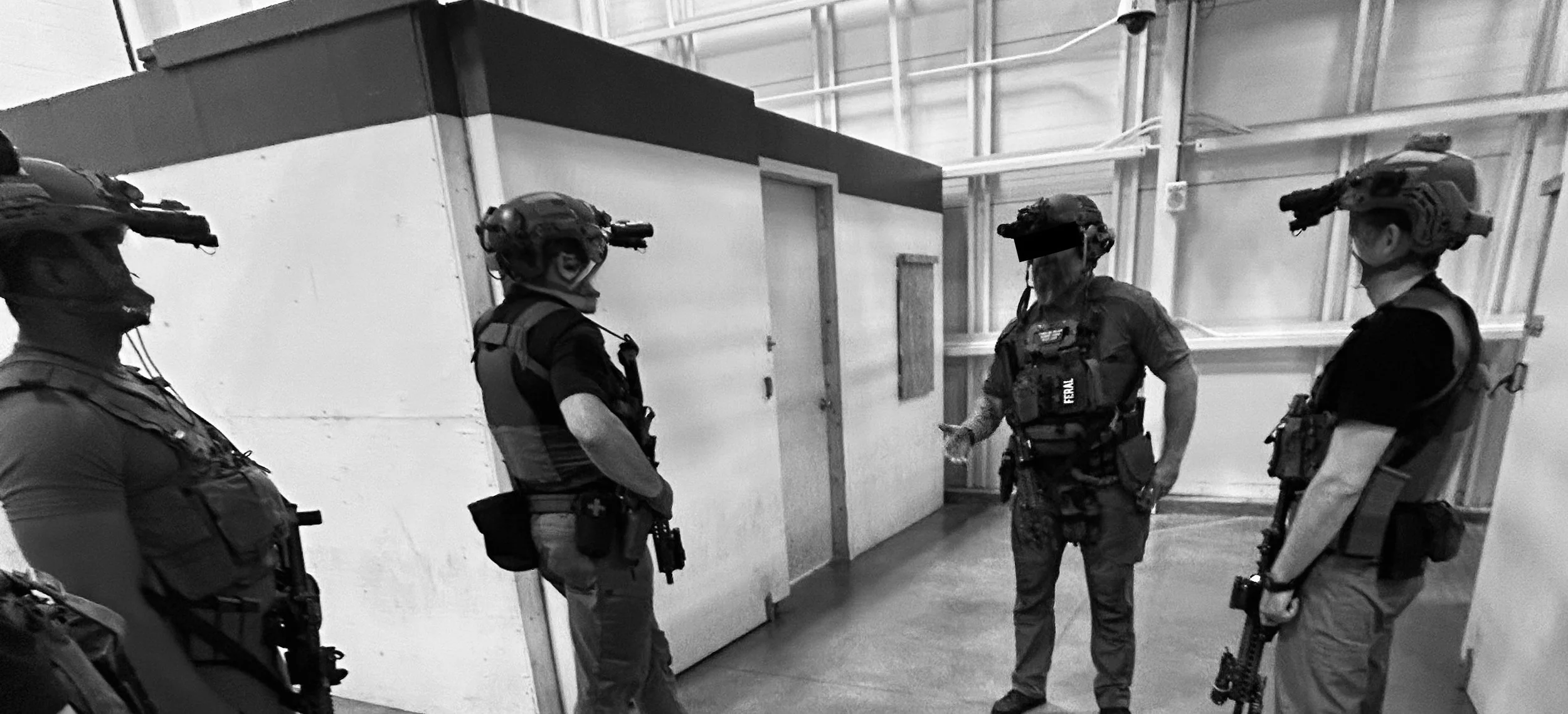 Four Security personnel in tactical gear standing in a warehouse-like indoor setting, appearing to be in a briefing or discussion.