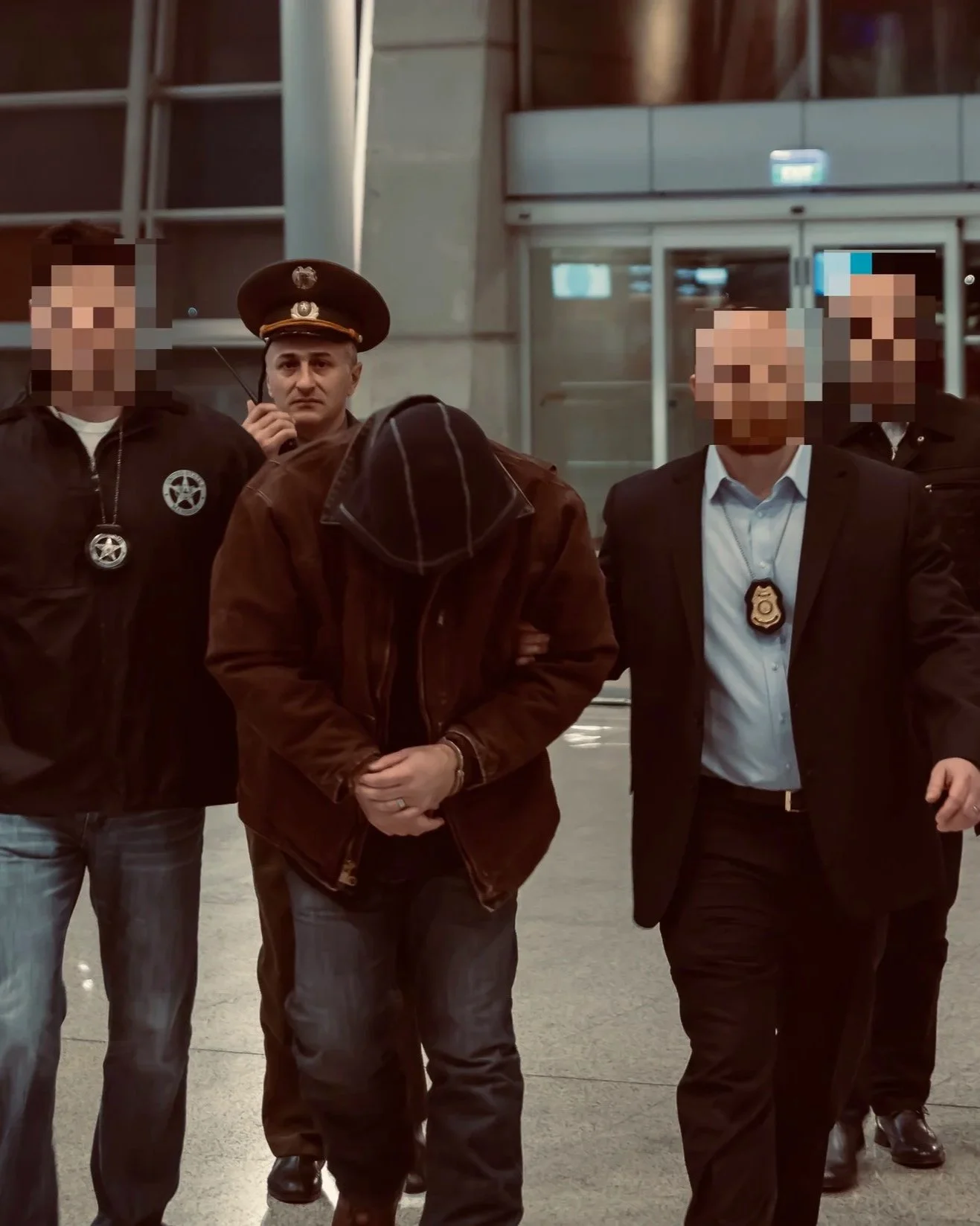 A man with his hands cuffed, being escorted by police officers through an airport or similar facility. The officers appear to be in plain clothes with badges visible, and the scene is indoors with metallic and glass architectural elements.
