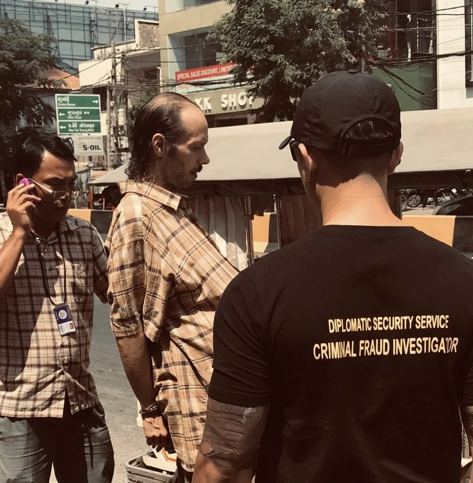 A man wearing a black shirt with the words 'Diplomatic Security Service Criminal Fraud Investigator' on the back stands in front of another man with long hair and a checkered shirt. A third man with glasses and a checkered shirt is on the phone. They are outdoors in an urban area with buildings, trees, and electrical wires visible in the background.