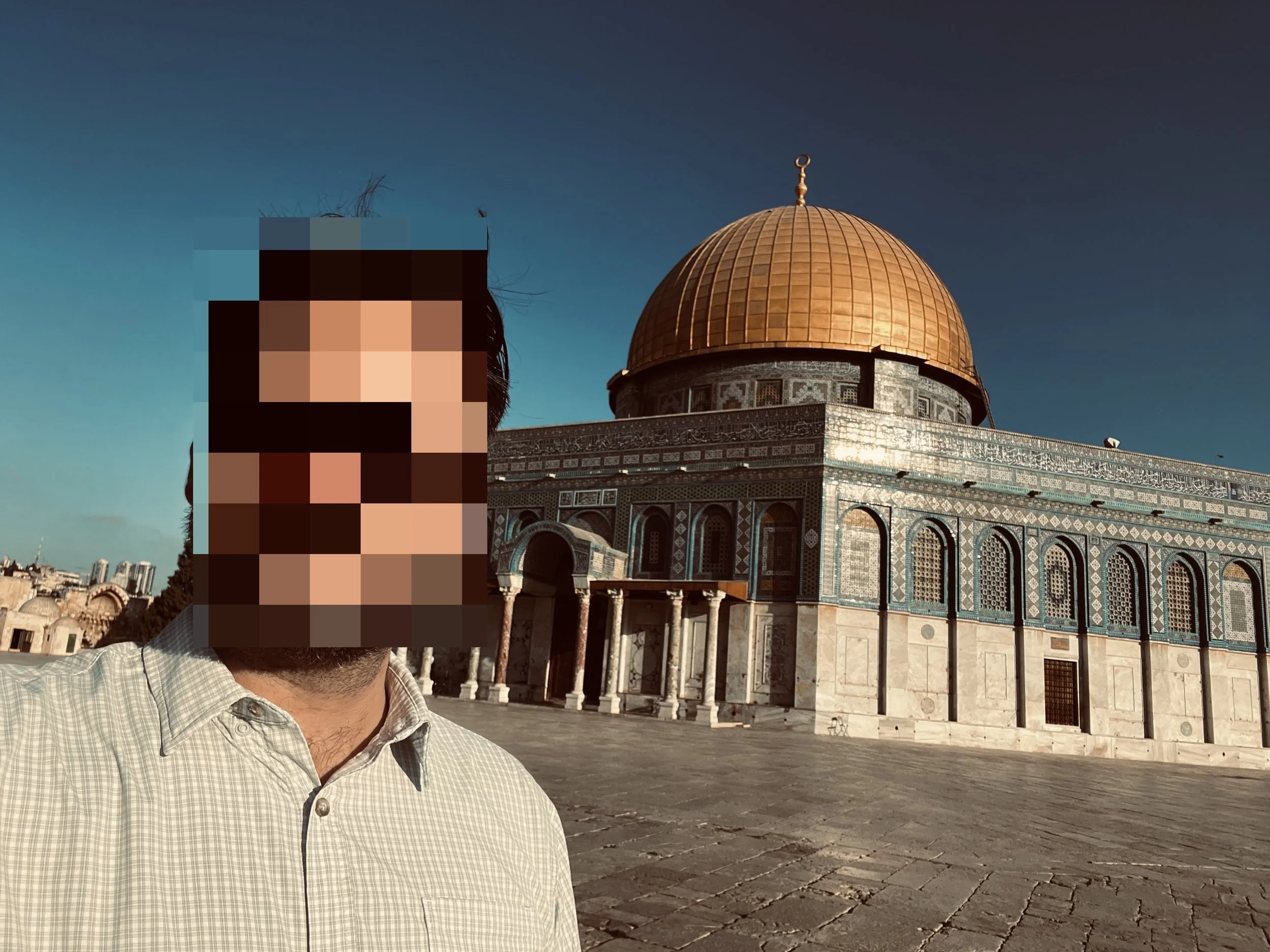 A person with a pixelated face wearing a light-colored checkered shirt, standing in front of the Dome of the Rock, a historic Islamic shrine with a large gold dome, located outdoors on a clear day.