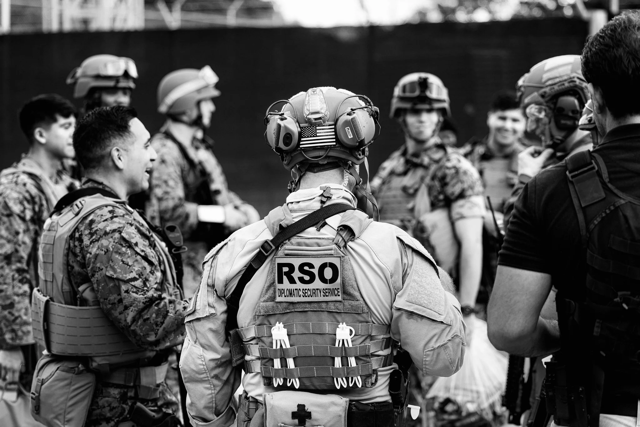 A group of military personnel and security officers gathered outdoors, some wearing helmets with visors and tactical gear, engaged in conversation.