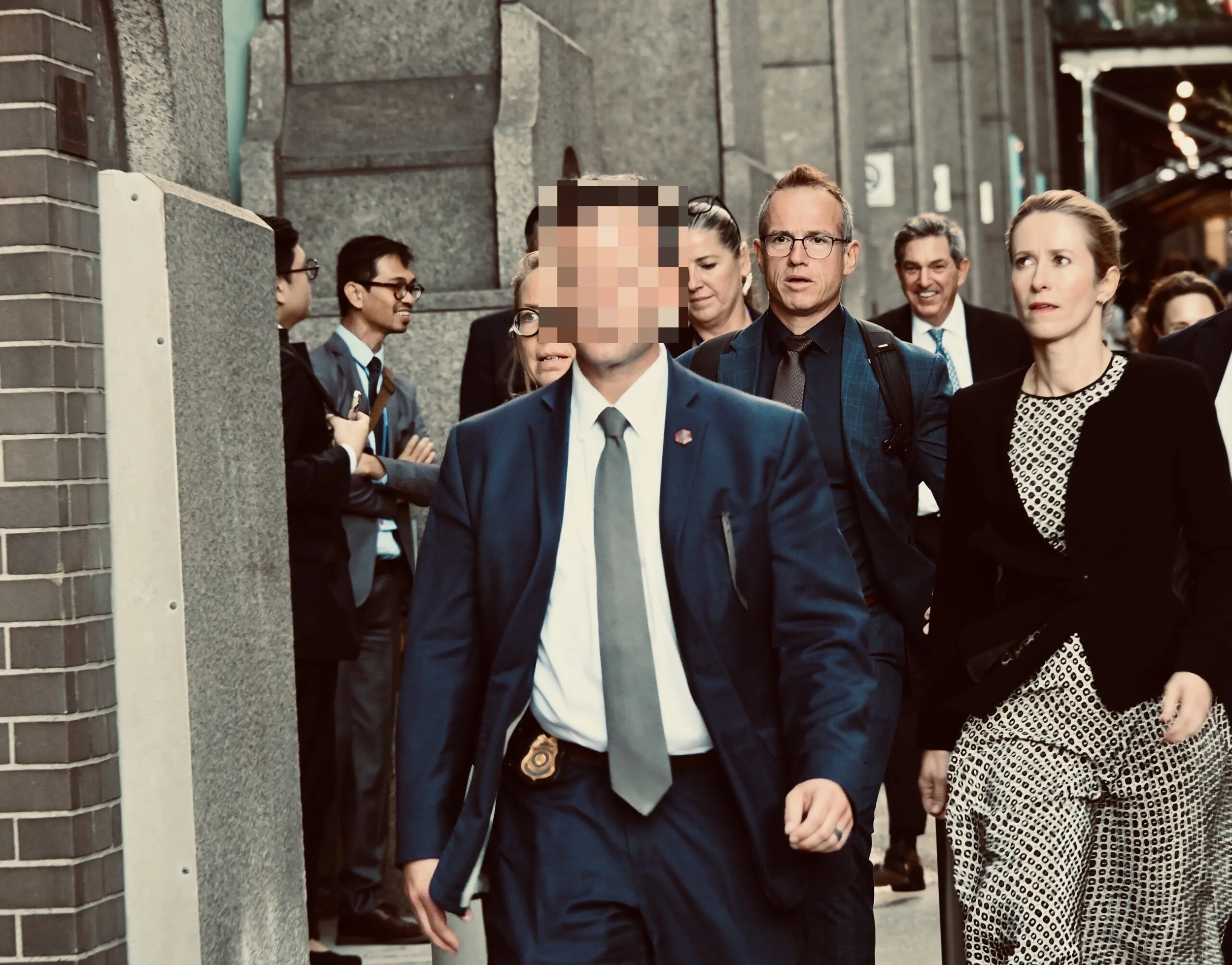 A group of professionally dressed people walking outside, with a man in the front wearing a suit and a badge on his belt. Others are following behind, including a woman in a black blazer and patterned pants, and a man with glasses and a backpack.