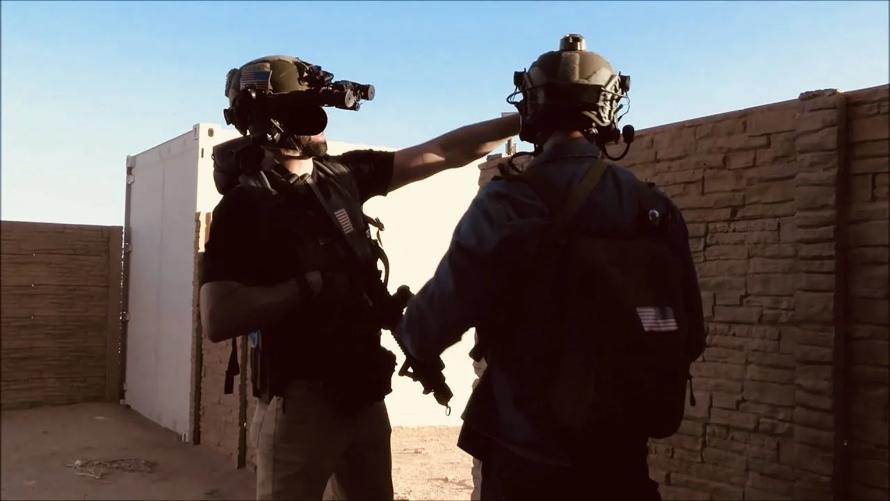 Two soldiers in tactical gear and helmets having a discussion outdoors near a brick wall under a clear sky.