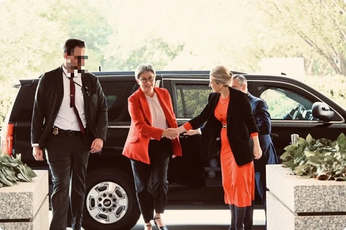 Two women shaking hands in front of a black SUV, while others look on, outdoors, with potted plants nearby.