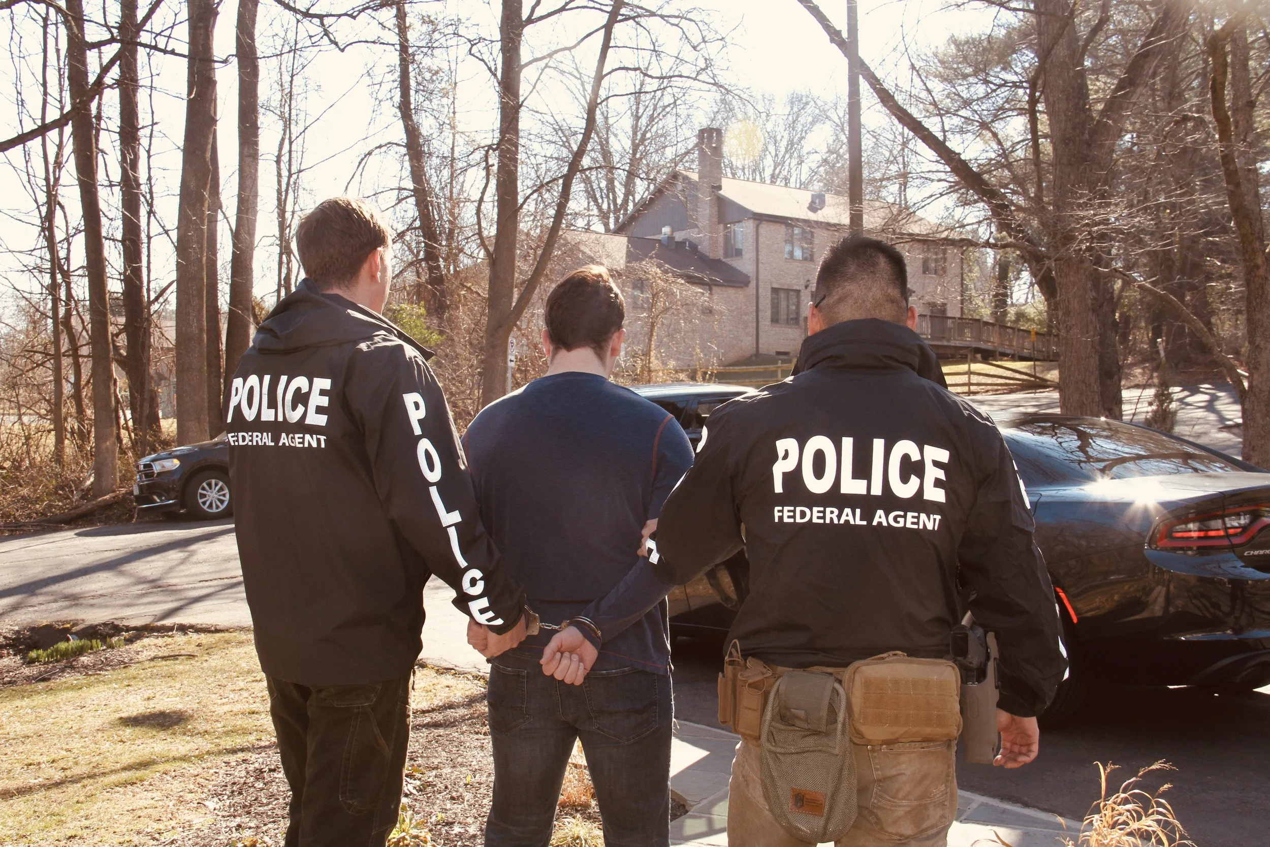 Two police officers in black jackets marked "POLICE FEDERAL AGENT" are handcuffing a man in a blue shirt on a sidewalk in a residential neighborhood. There are trees and houses in the background.