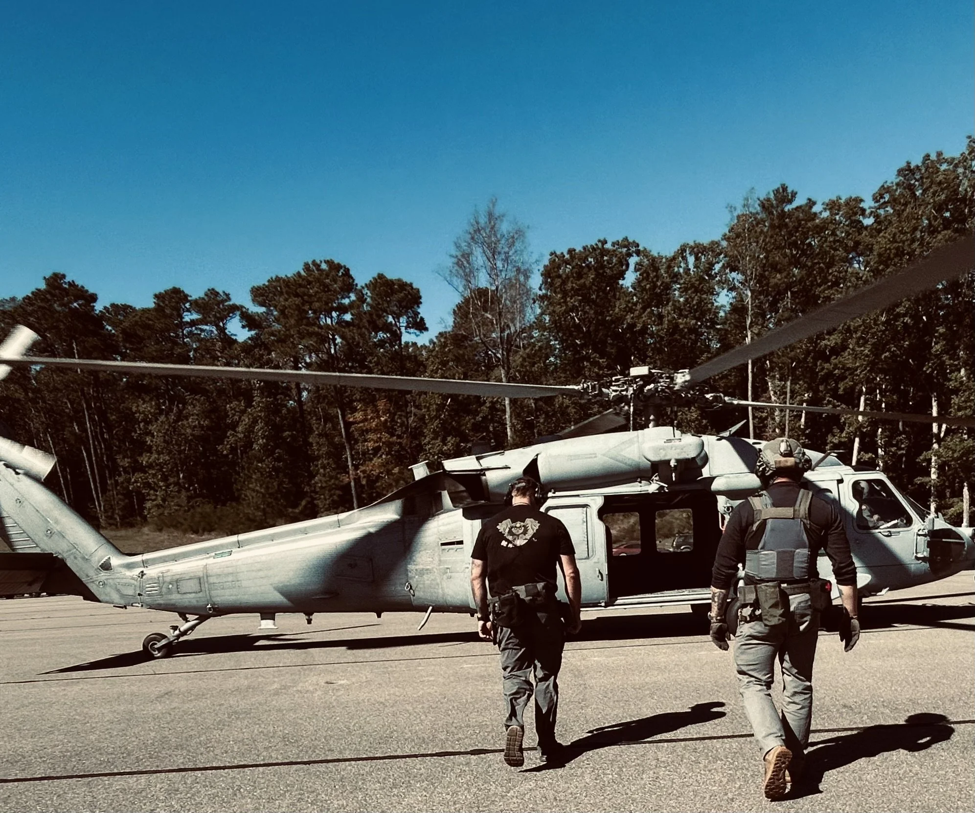 Two military personnel walking towards a gray military helicopter on a tarmac, with trees and a clear blue sky in the background.