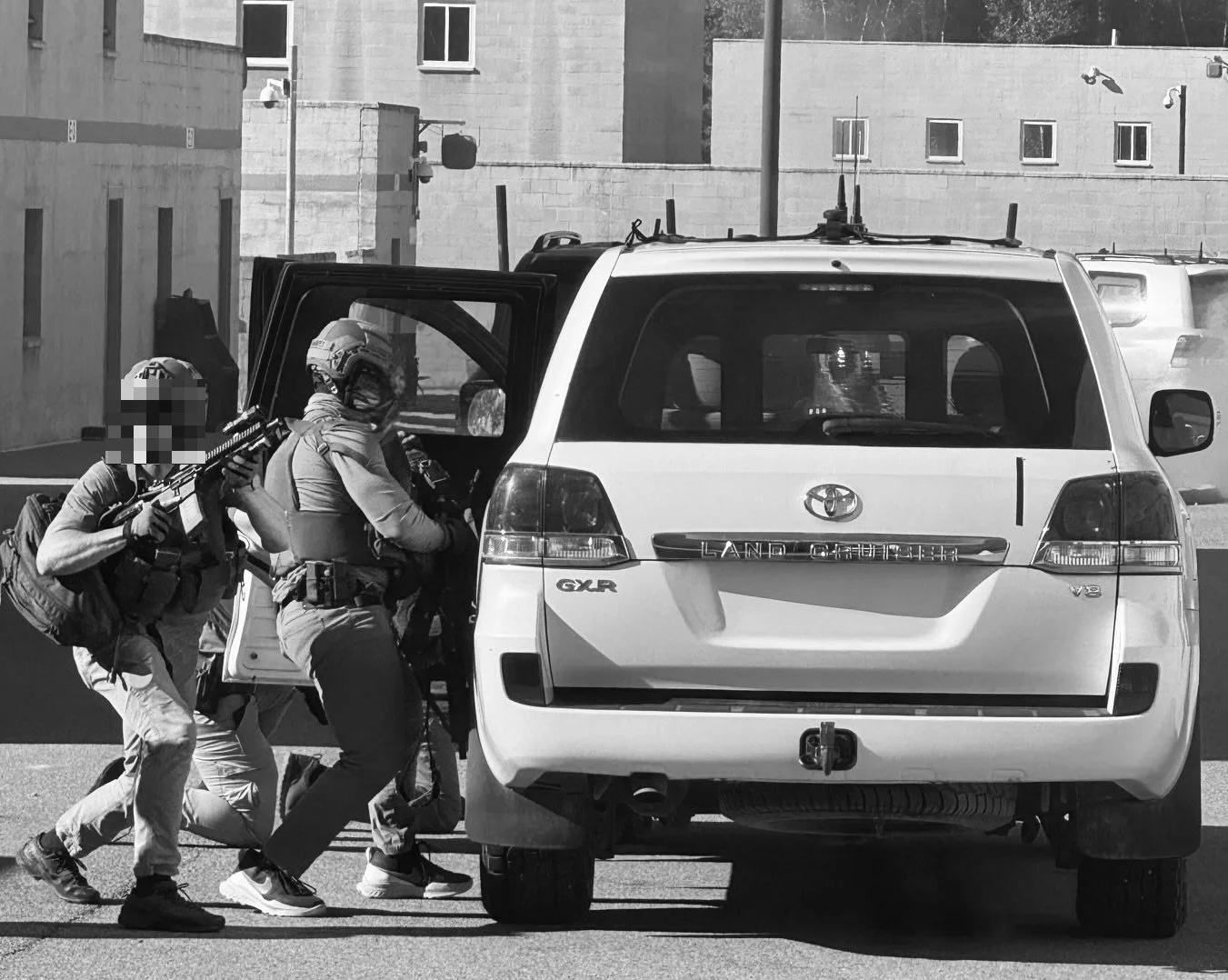 Two armed tactical officers with face masks and helmets are approaching the open door of a white Toyota Land Cruiser SUV in an urban area. One officer is holding a rifle, while the other appears to be preparing to enter the vehicle. The scene is in black and white.
