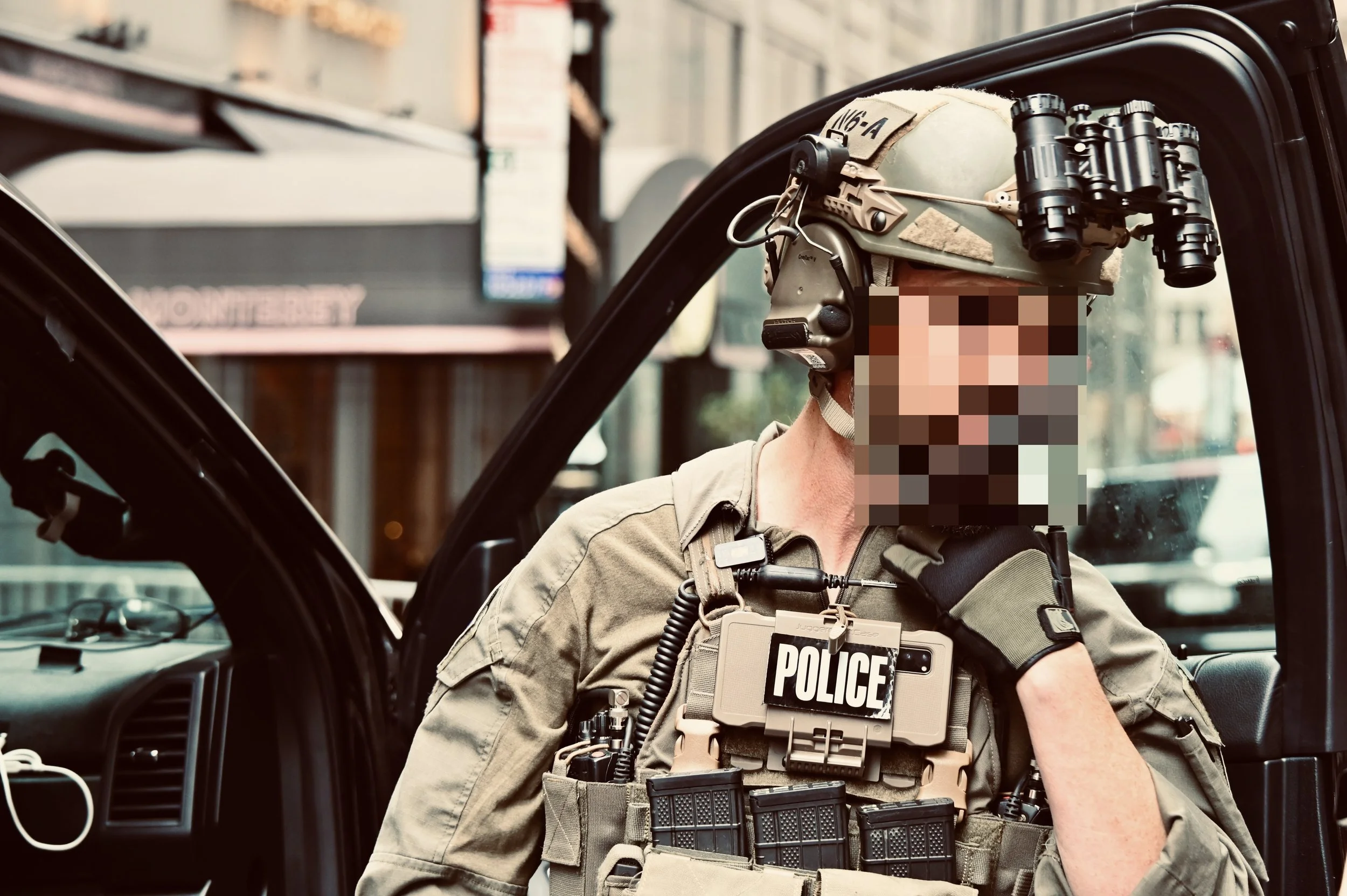 A police officer wearing tactical gear and a helmet with night vision goggles, sitting inside a vehicle with the door open, in an urban setting.
