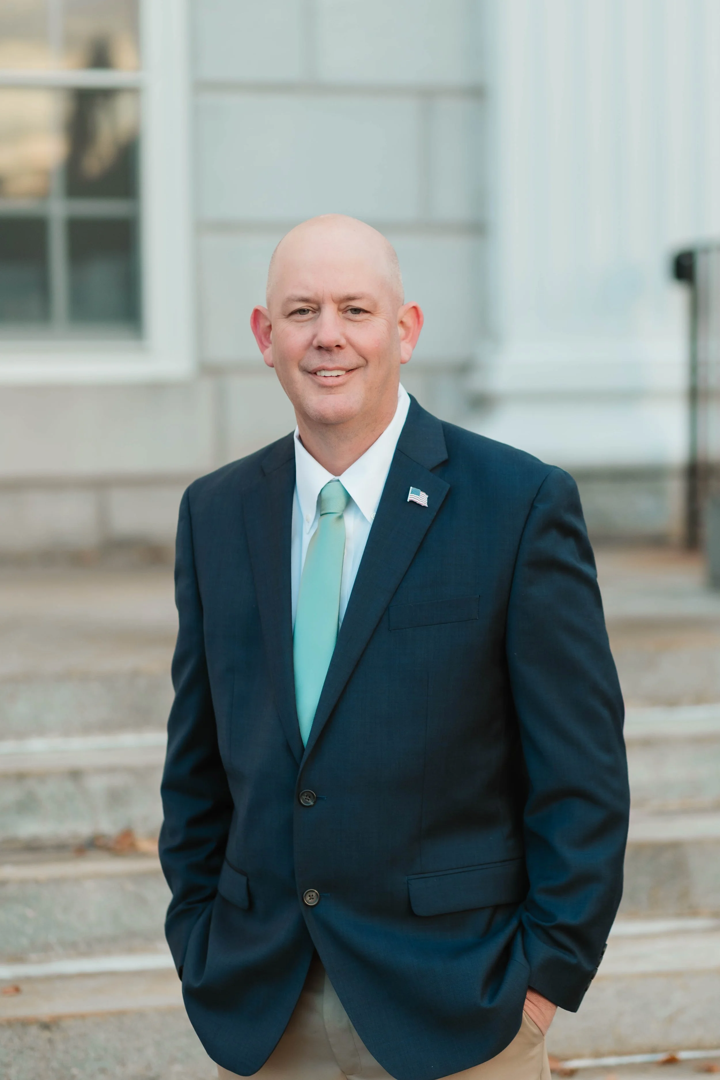 A man in a navy suit with a light blue tie and American flag lapel pin standing outdoors in front of a building with steps.